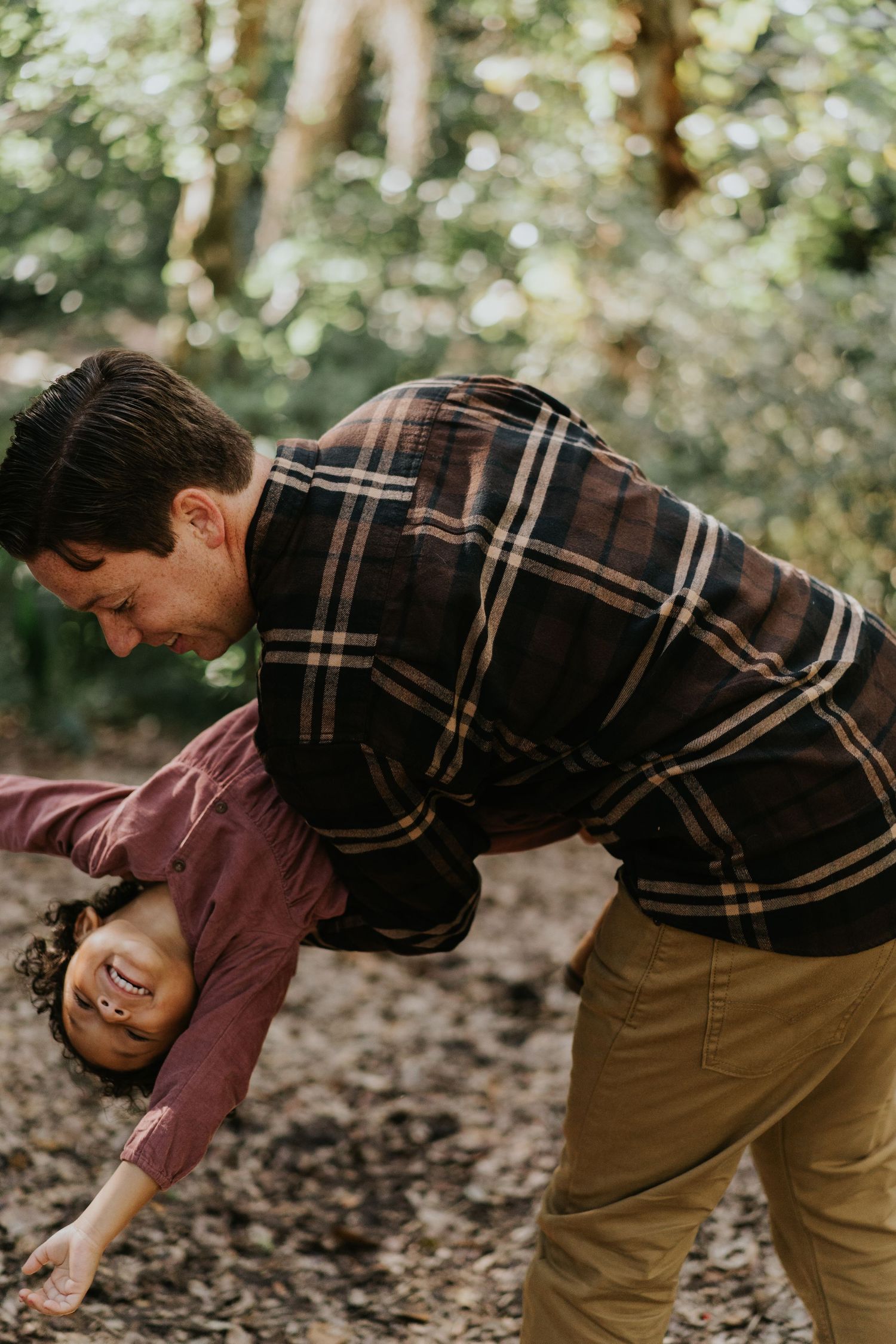 A couple shares a playful moment laughing together in a forest setting with warm sunlight filtering through trees.