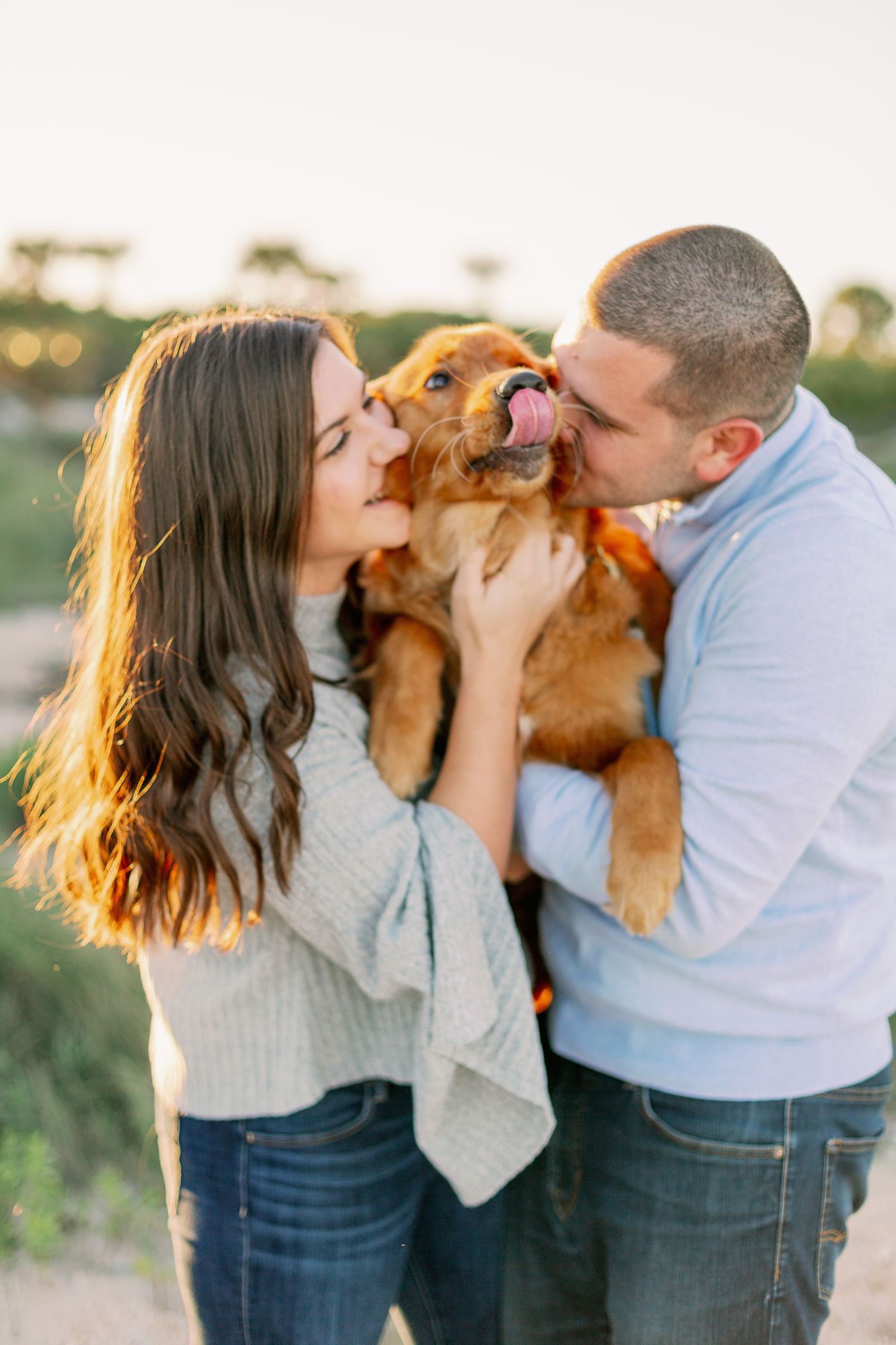 A couple shares affectionate moments with their brown dog in a sunny outdoor setting.