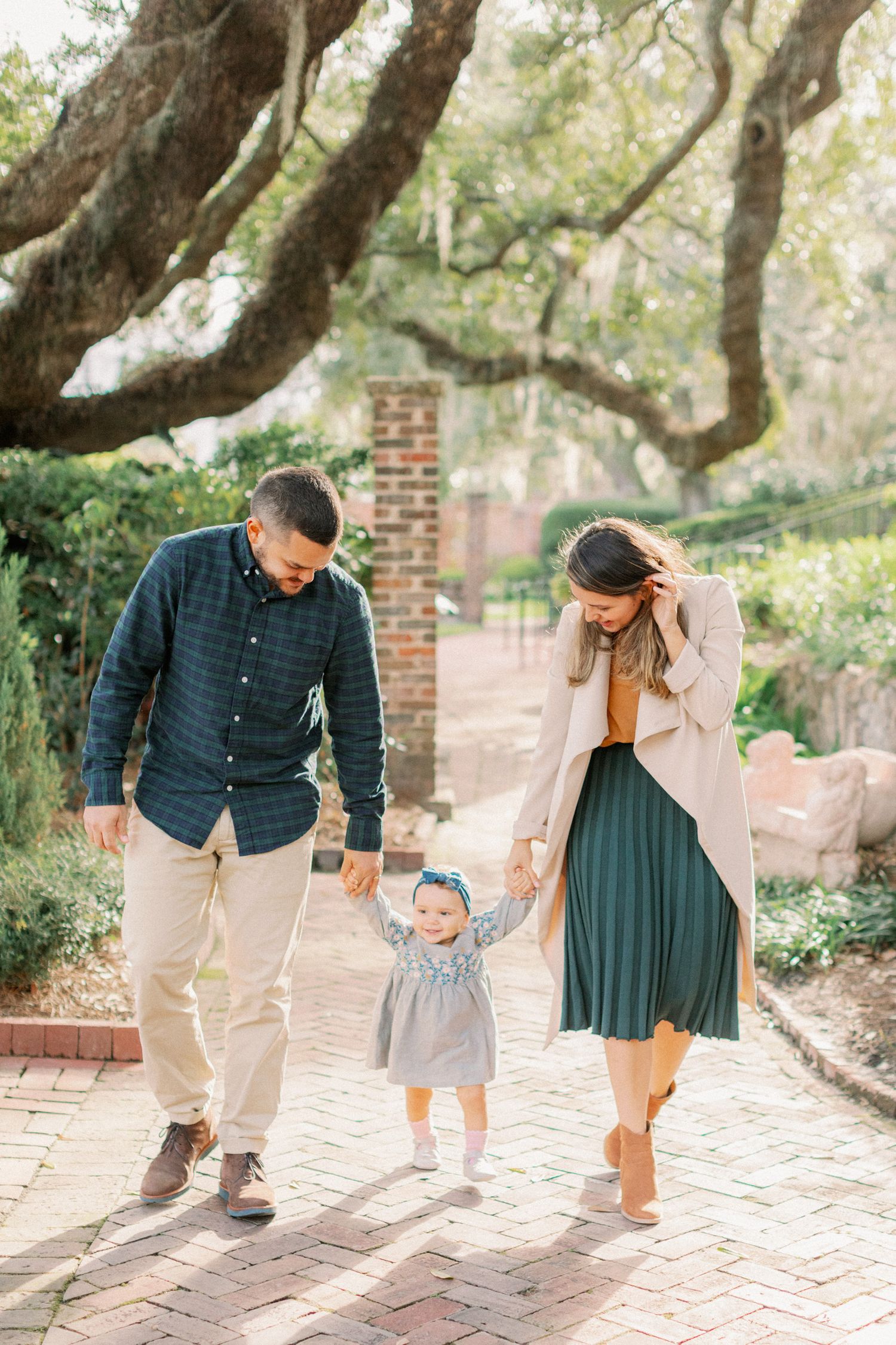 A family walking and playing together along a brick pathway under large oak trees.