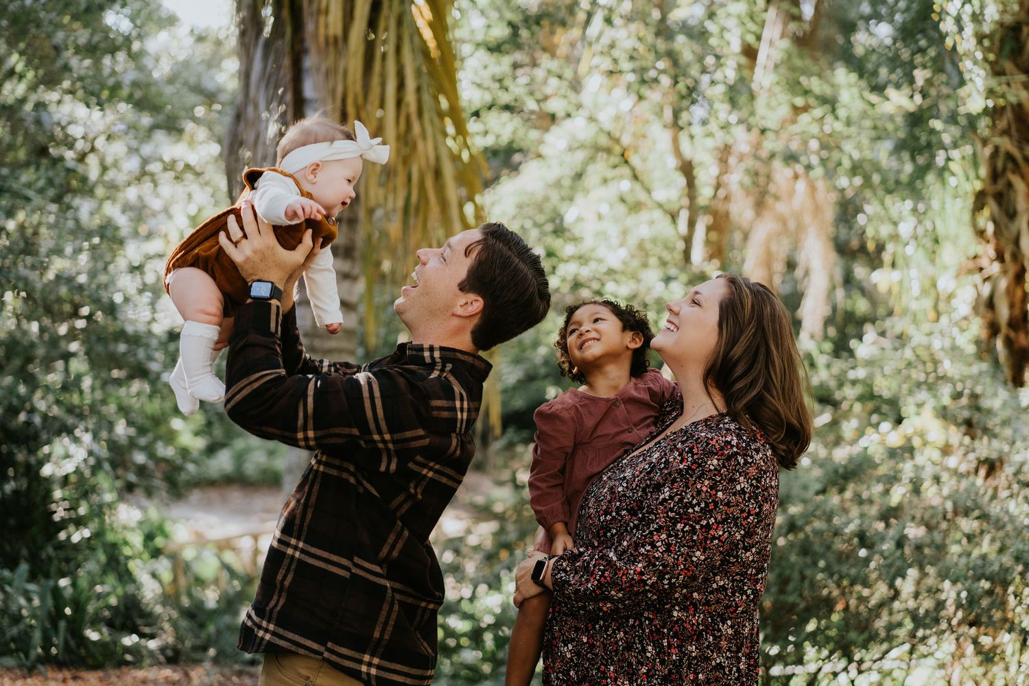 A family enjoys a playful moment together in a sunlit forest setting, lifting a baby up in the air.