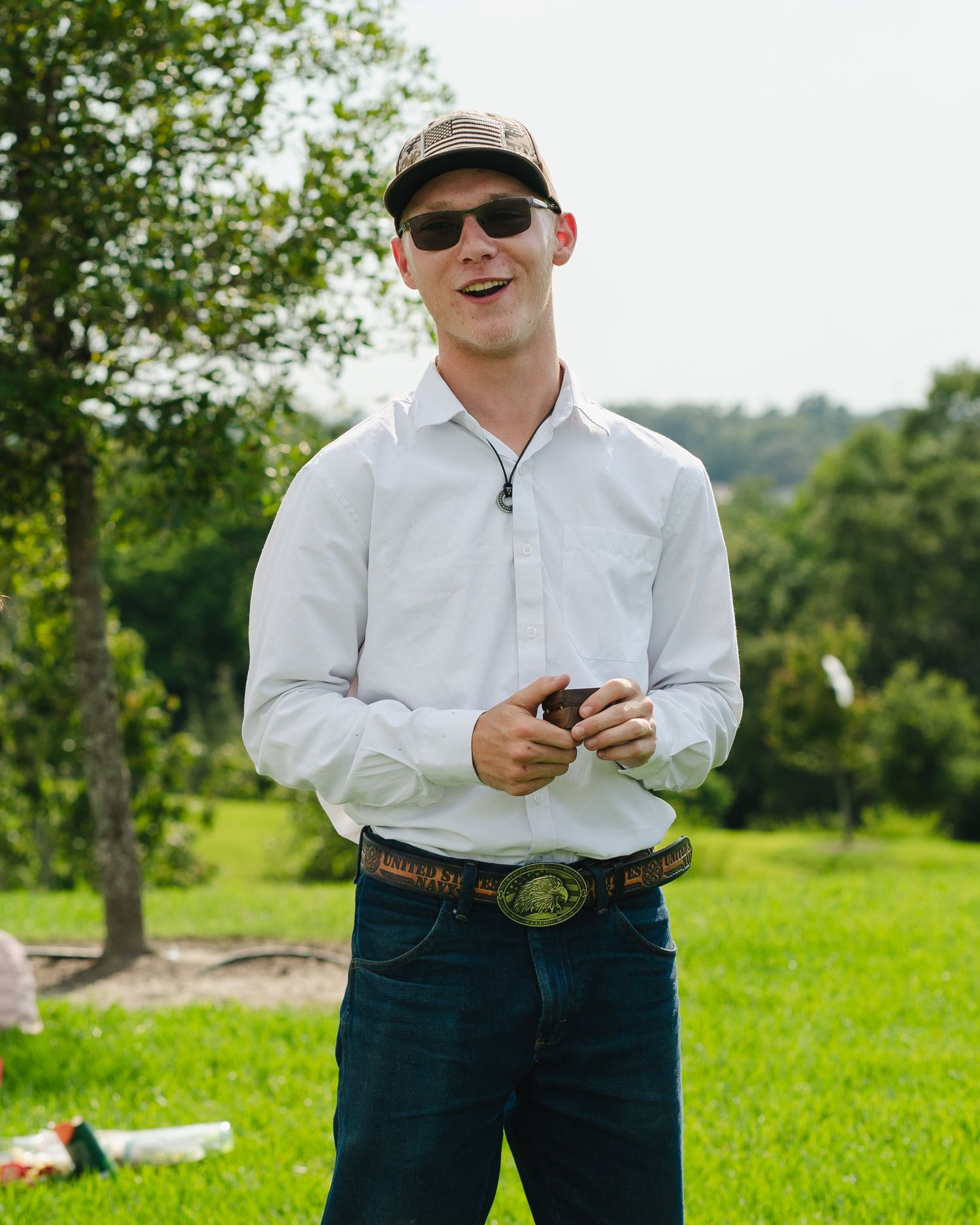 A person in sunglasses and white shirt stands in a grassy field holding something.