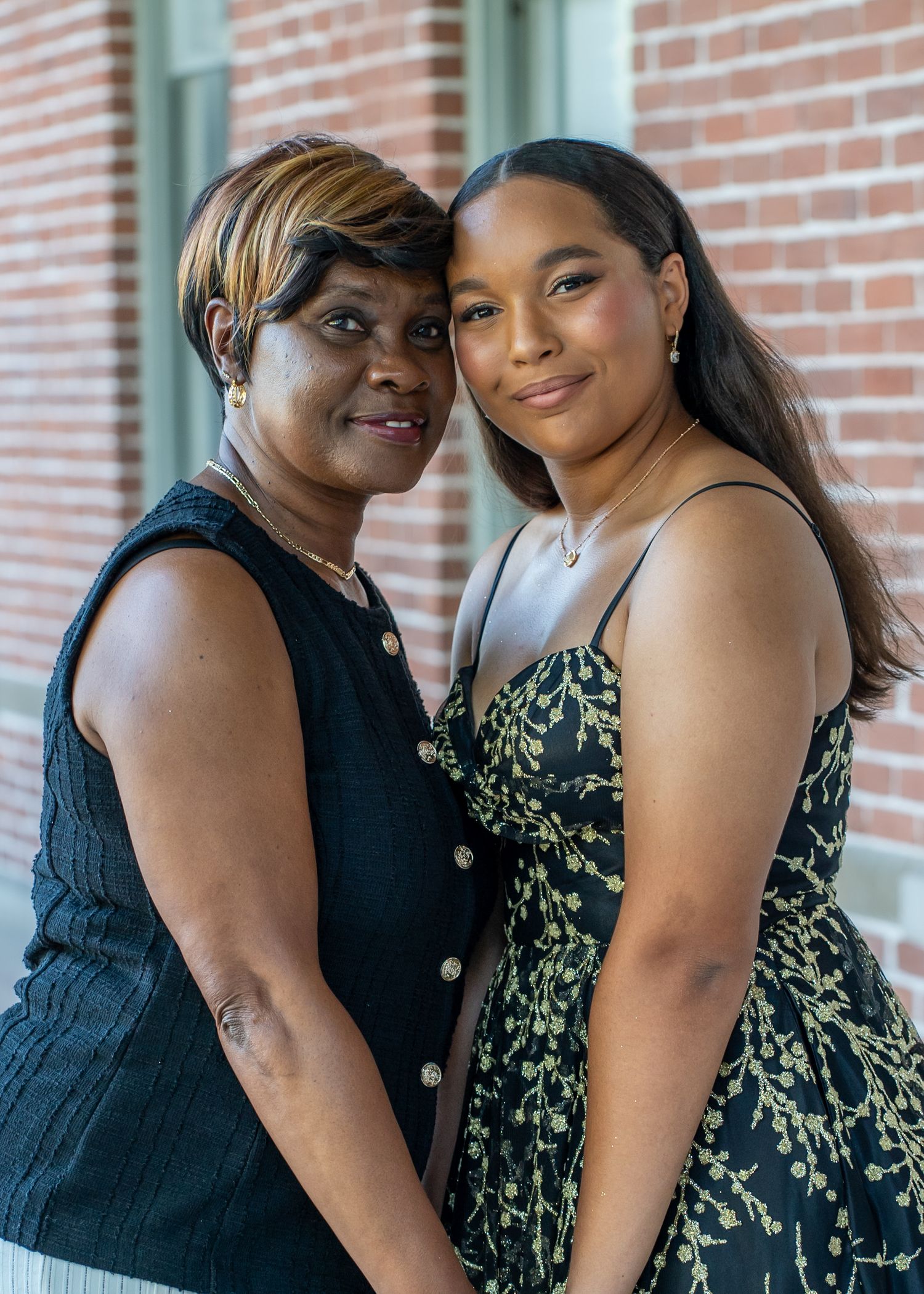 Two people pose together in elegant attire against a brick wall backdrop.