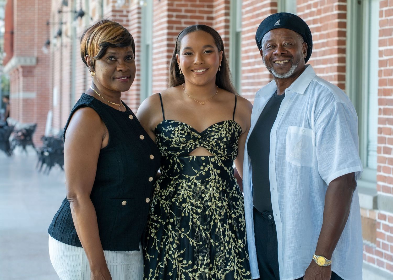 Three people pose together on a brick-lined porch in casual summer attire.