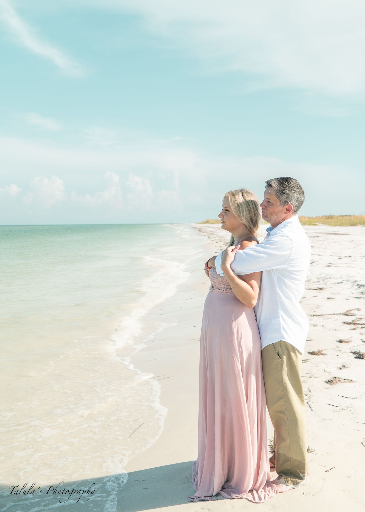A couple embraces on a sunny beach with white sand and turquoise waters in the background.