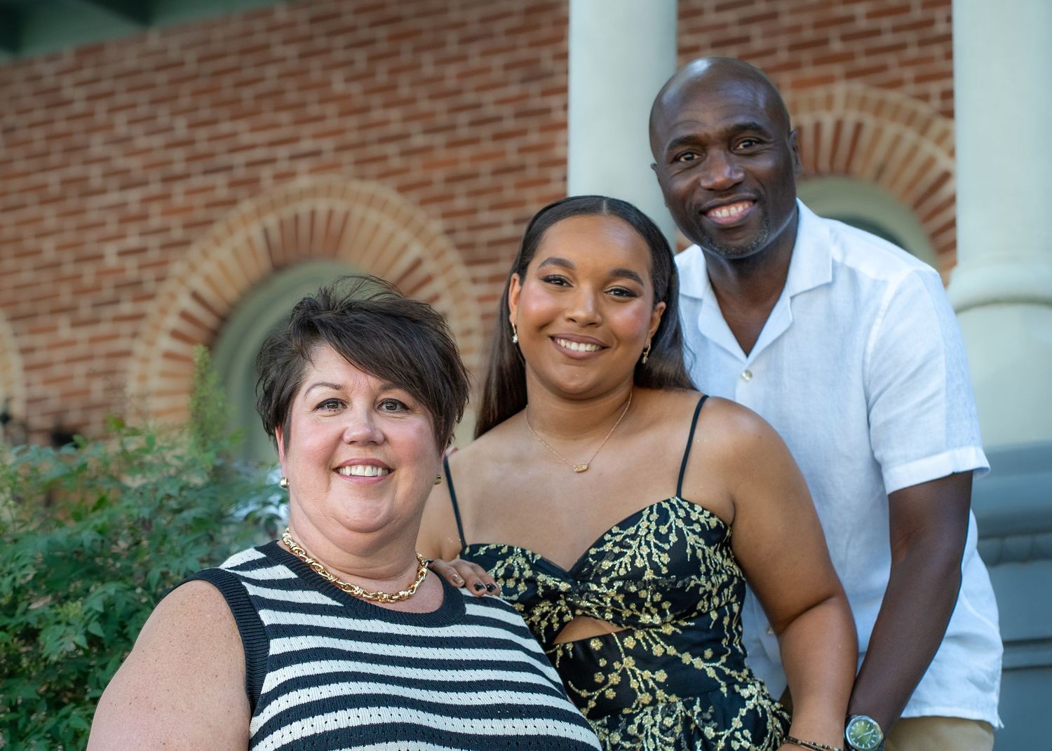 Three people smile together on a porch with brick and column architecture.