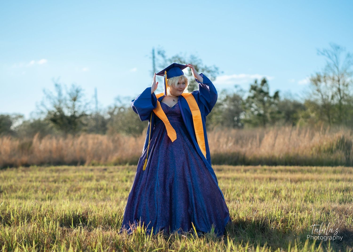 Graduate wearing blue cap and gown poses in a sunny outdoor field setting at sunset.