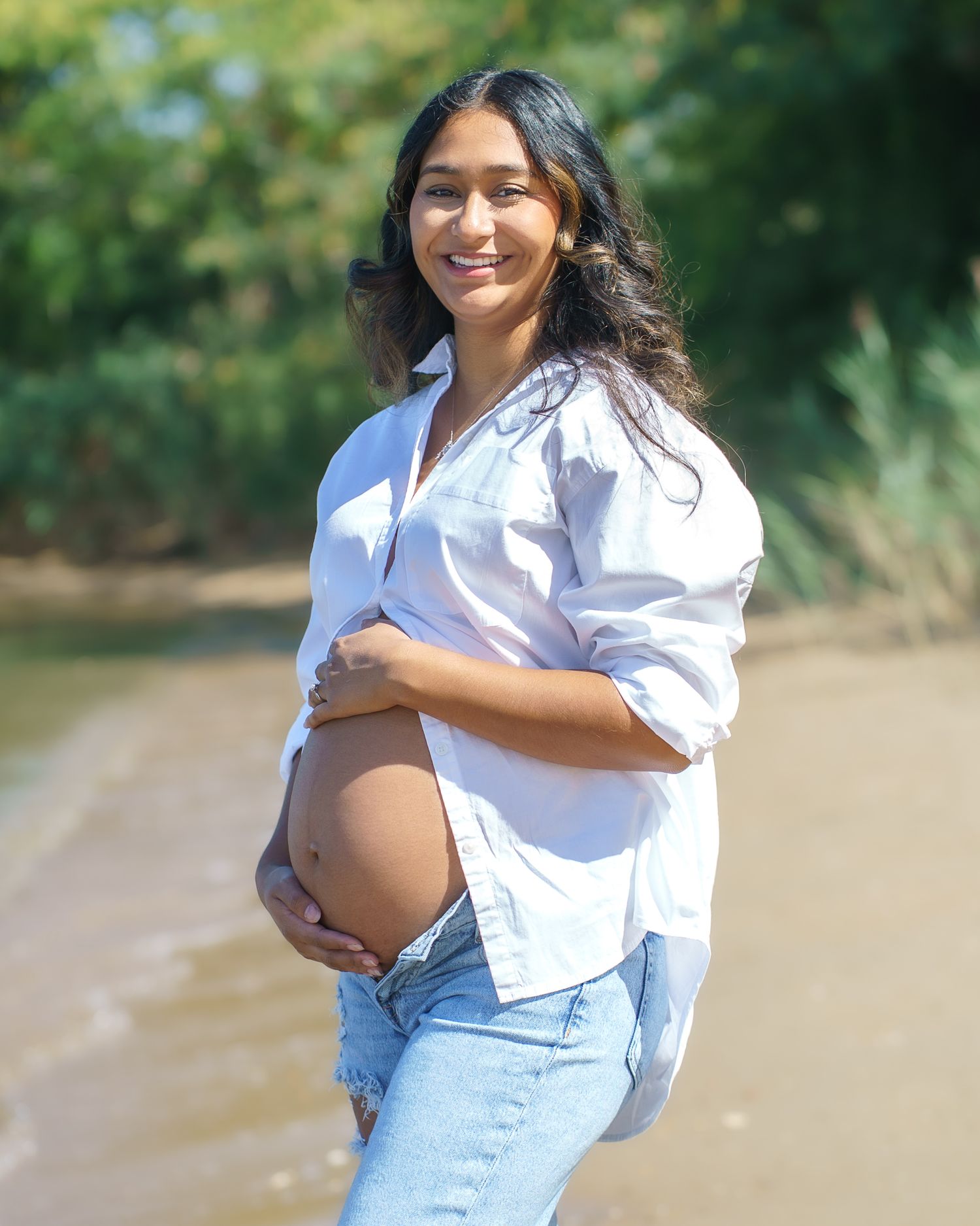 A maternity photoshoot featuring a white shirt and jeans against greenery.