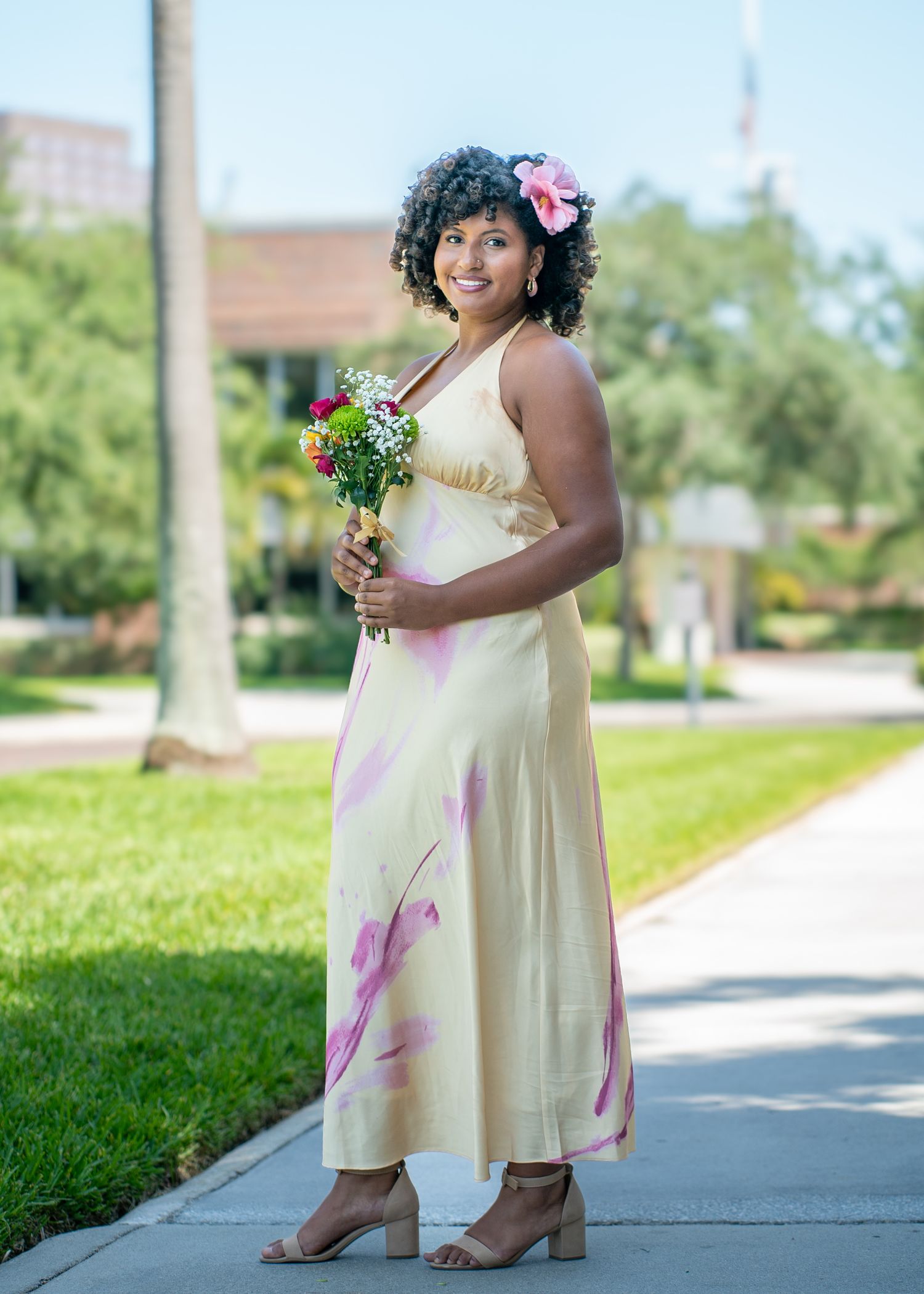 A person in a cream dress with pink accents holds flowers while standing on a sidewalk.