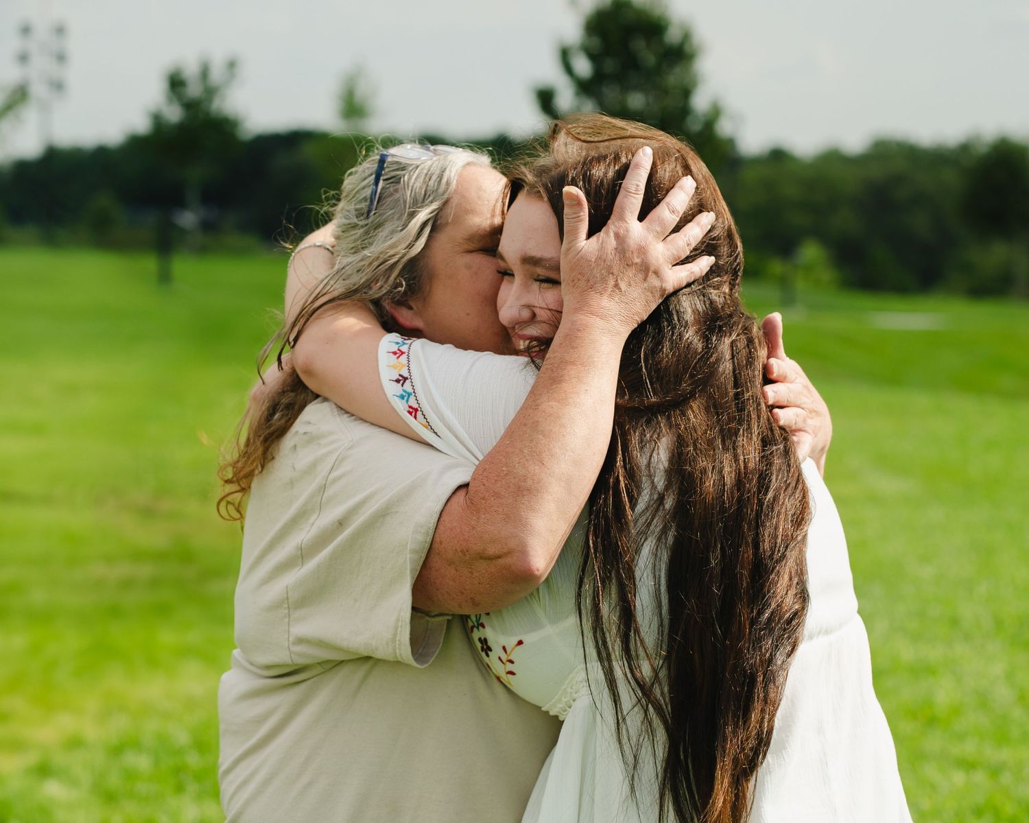 A couple shares an intimate moment in a sunny field with trees in the background.