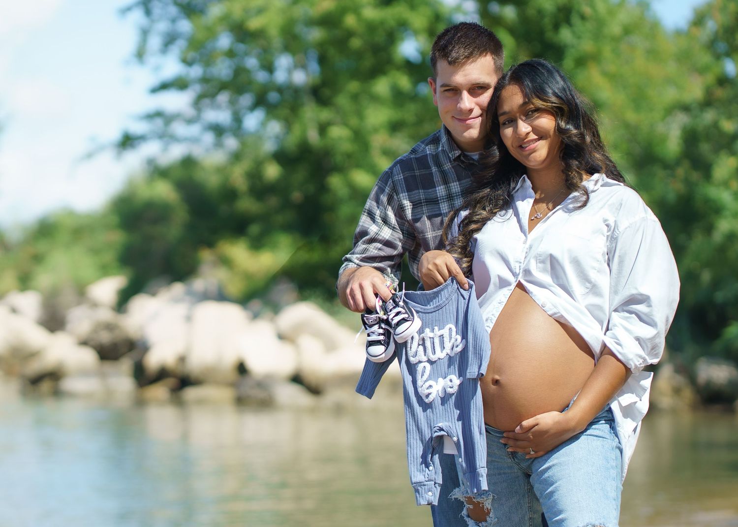 Expectant couple showing baby clothes during outdoor photoshoot.