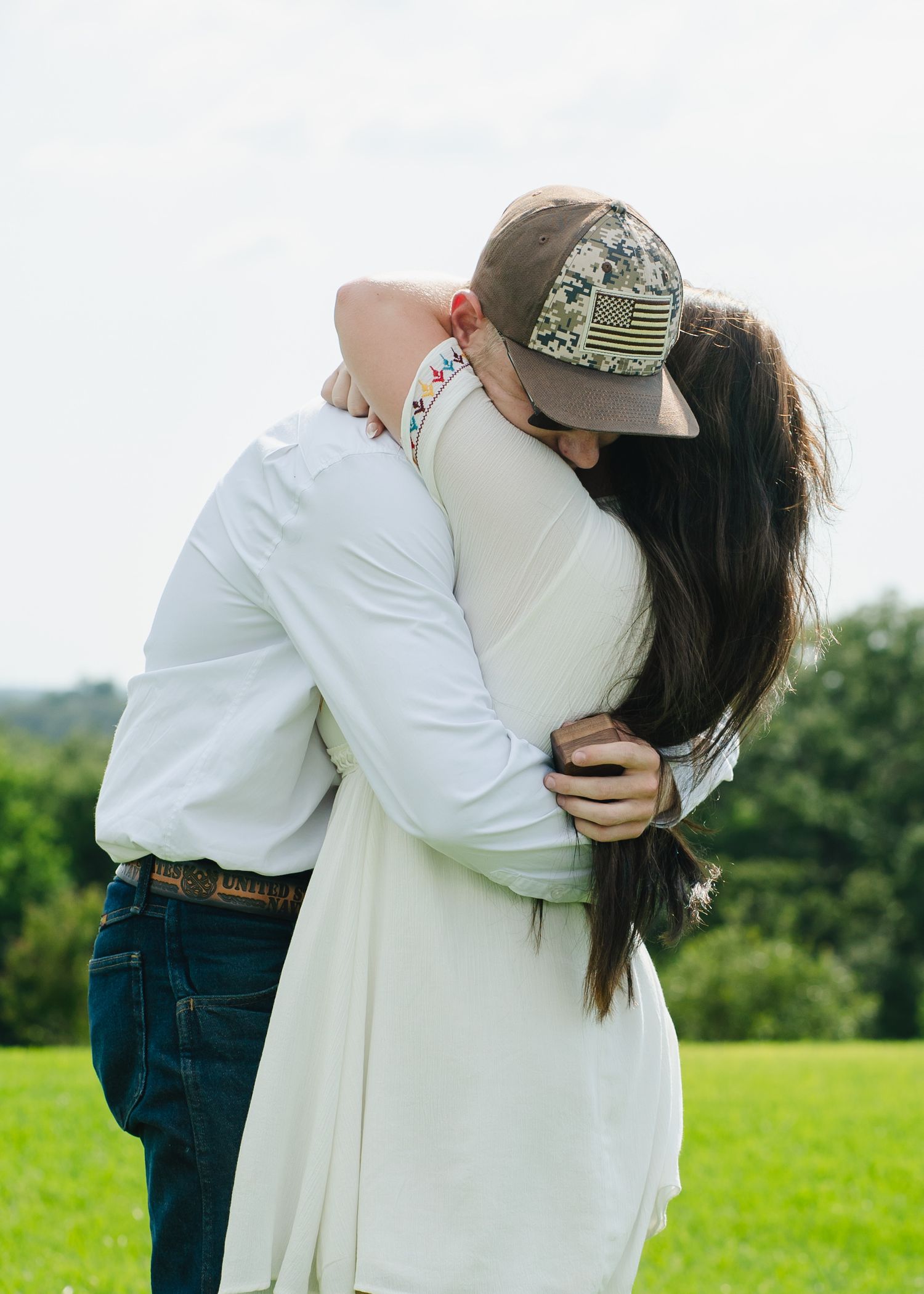 Two people embrace in a white dress and casual wear on a sunny green field.