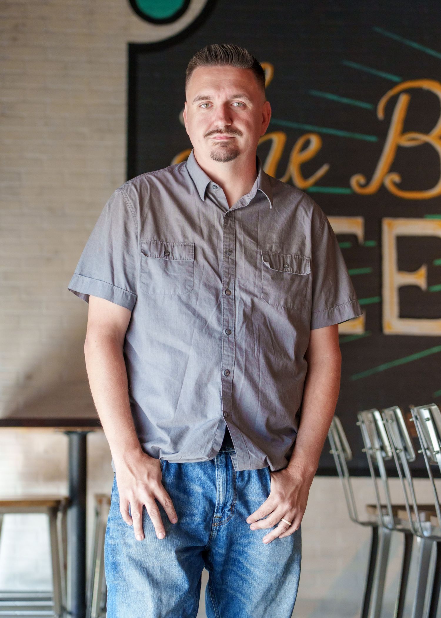 Man in gray button-down shirt and jeans posing in a casual restaurant setting.