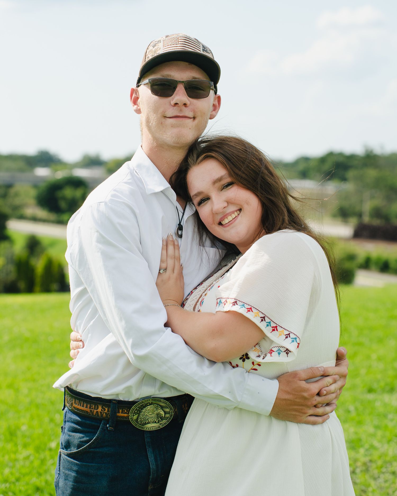 A couple embraces while wearing casual attire in a sunny green field.