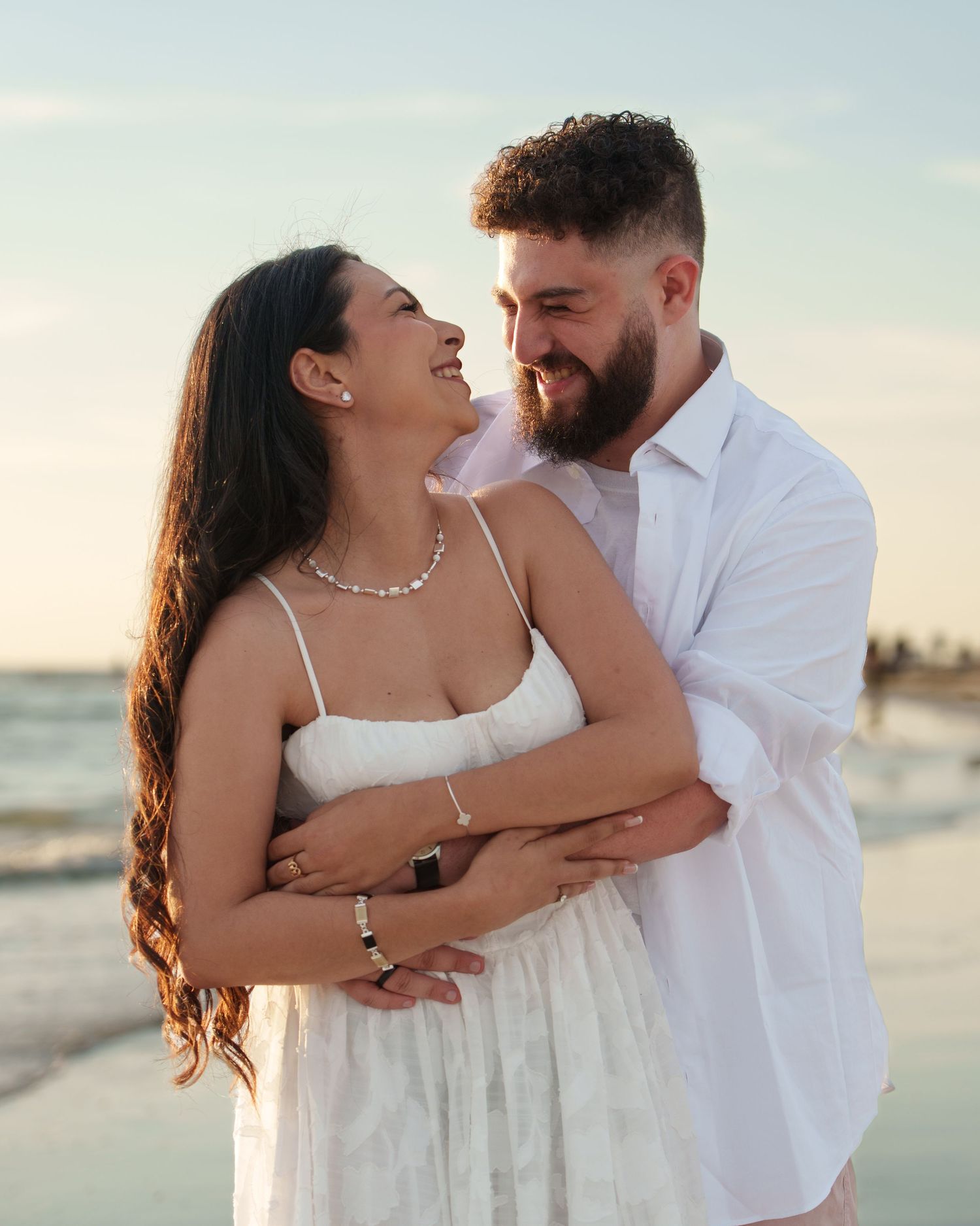 A couple shares a joyful moment together on the beach in white outfits.