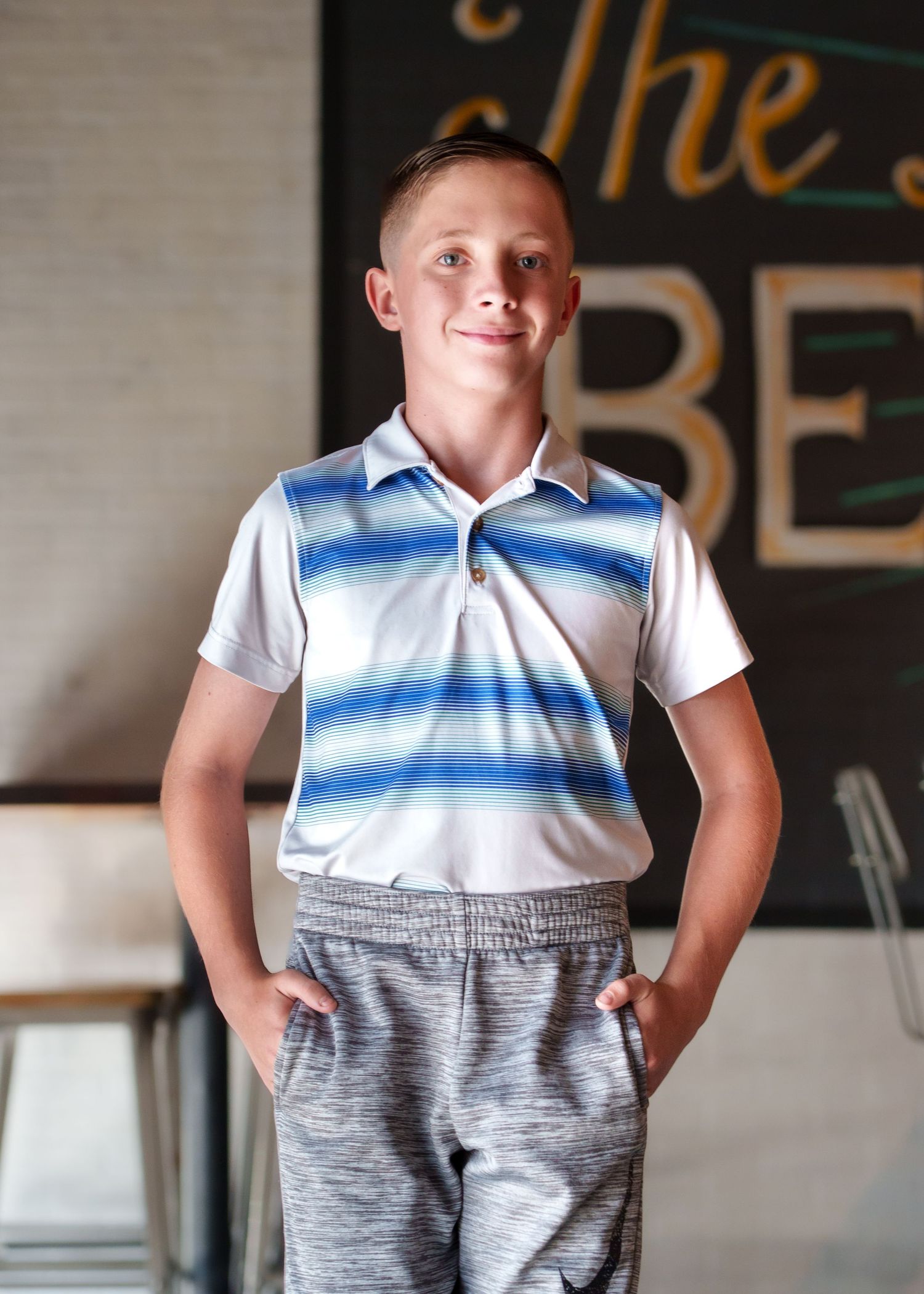 Child in striped polo and gray pants striking playful poses in restaurant setting.