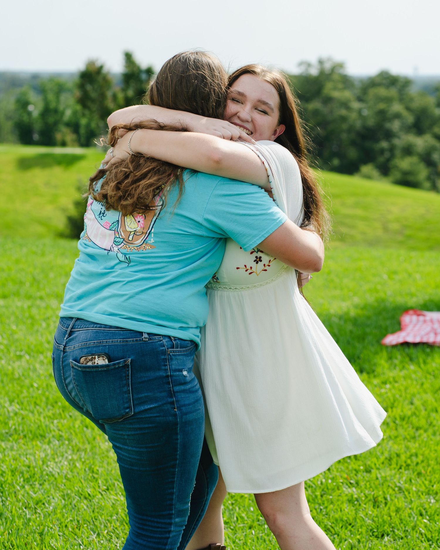 Two people embrace warmly on a green hillside during a sunny afternoon.