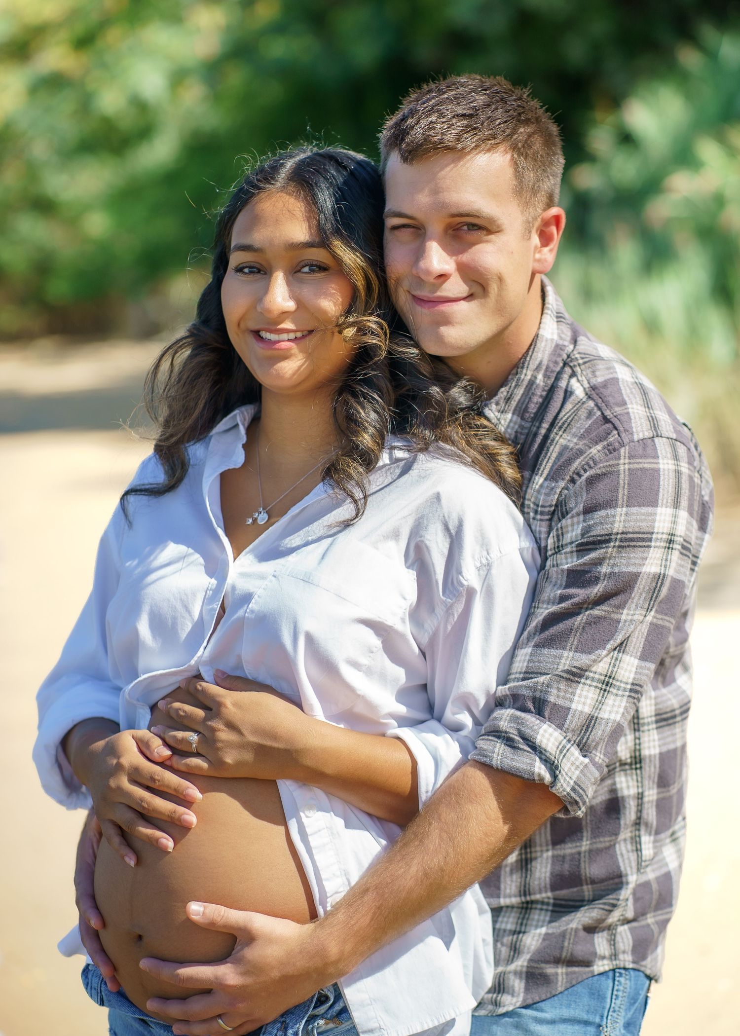 A couple embraces outdoors in casual attire against a natural green background.
