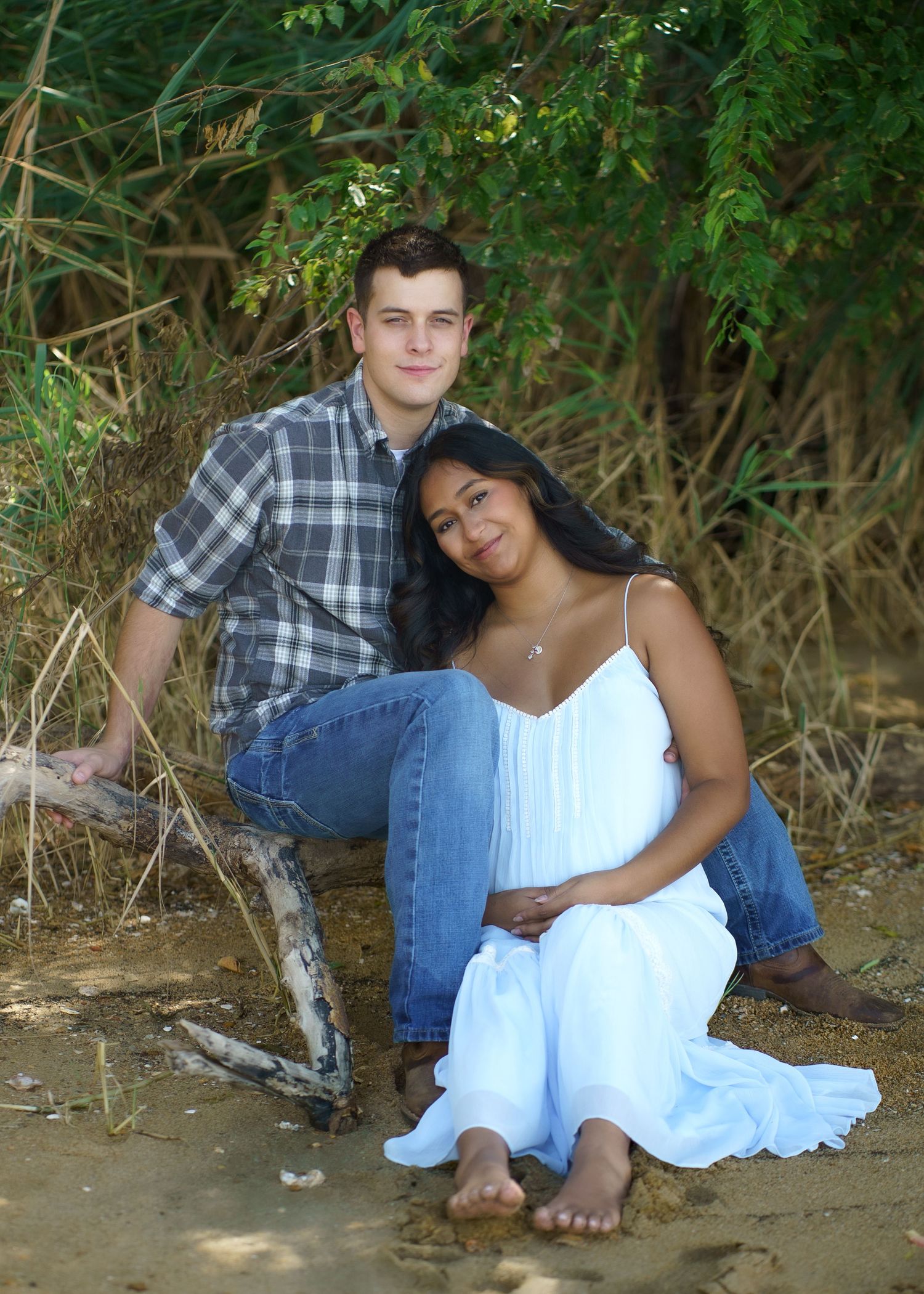A couple poses casually on fallen logs near a waterfront setting.