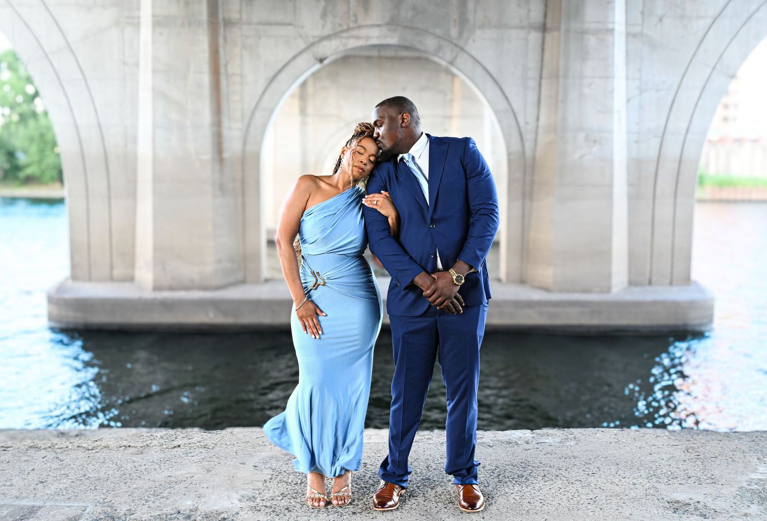 A couple poses under a bridge wearing formal blue and navy attire.