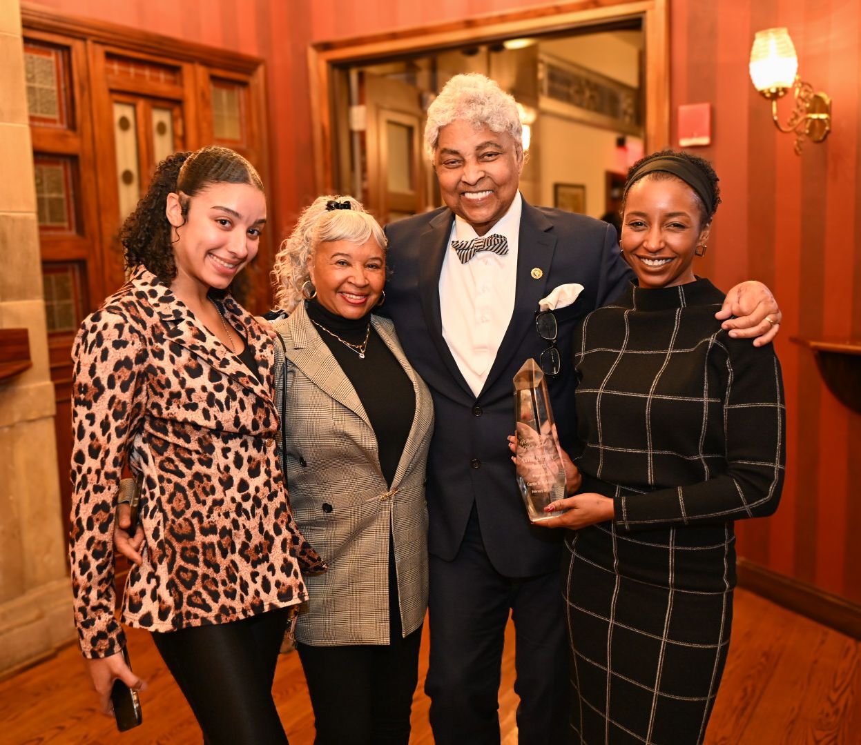 A group of four people pose together at a formal event, with one wearing a tuxedo and others in business attire.