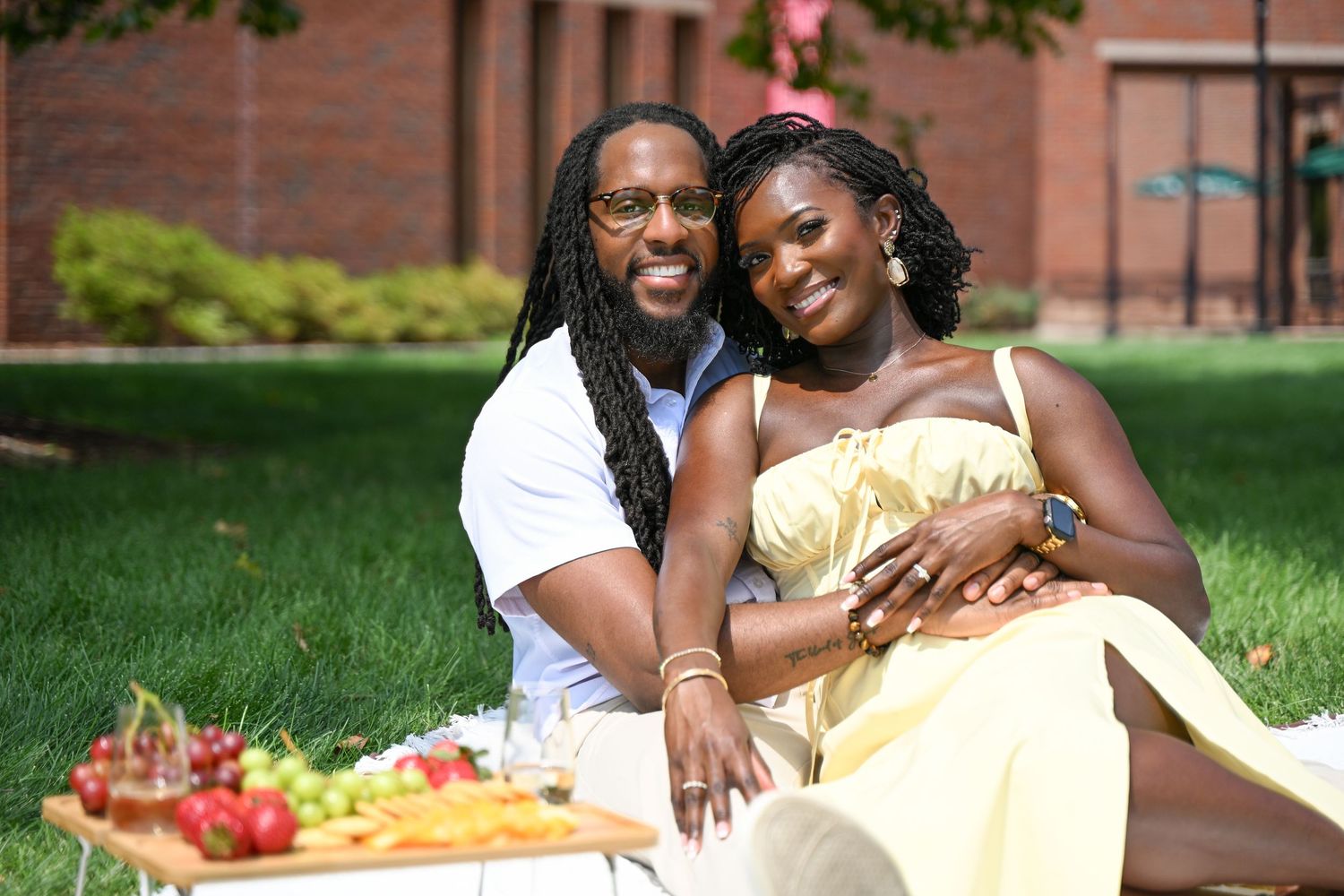 A couple enjoys a romantic outdoor picnic on a lawn with fresh fruits and snacks laid out on a blanket.