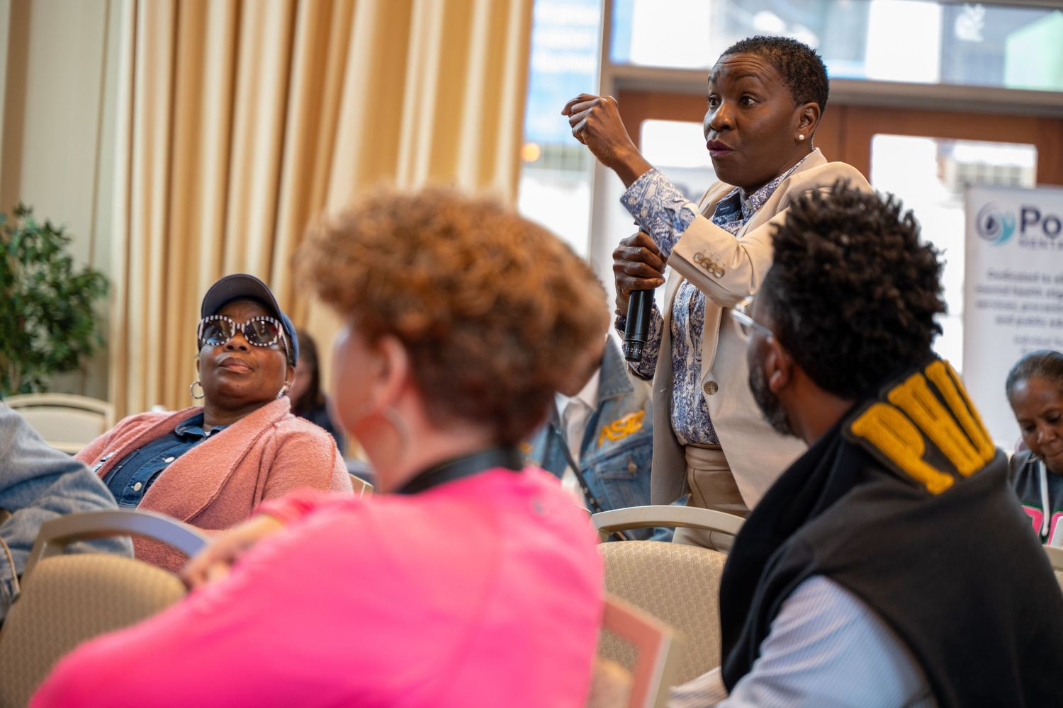 People engage in conversation at a community gathering with beige curtains and indoor lighting visible in the background.