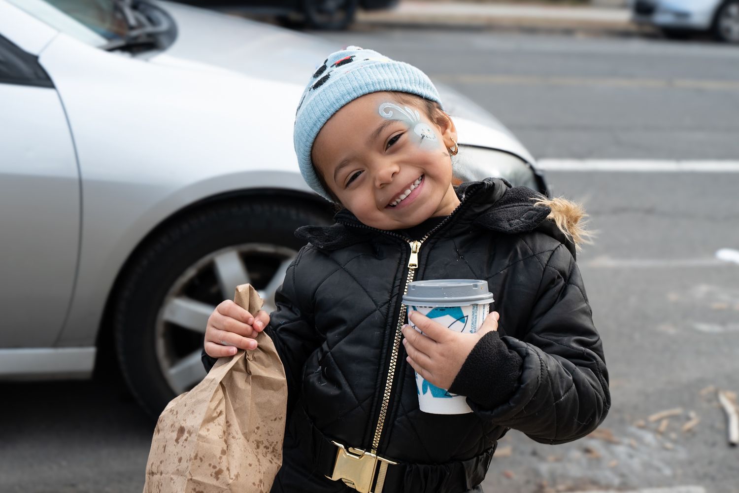 A person in a black winter coat holds a brown paper bag and coffee cup while standing on a city street.