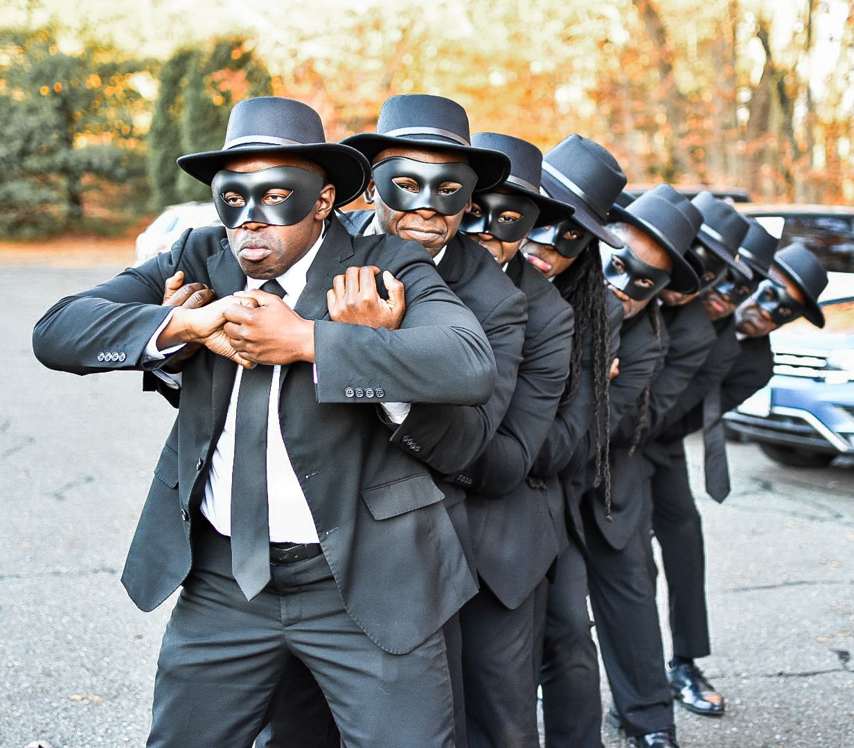 Men in matching black suits and hats striking synchronized poses outdoors.