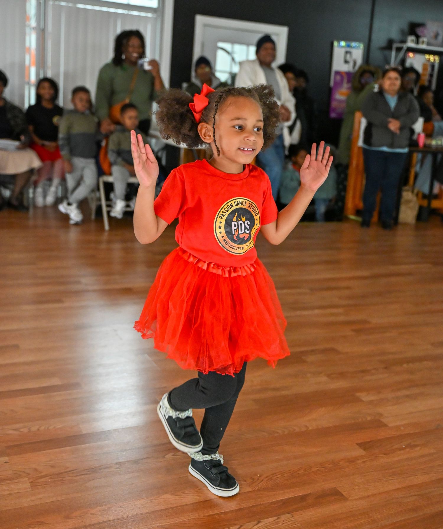 Young dancer in a bright red tutu performing energetic moves on a wooden dance floor.