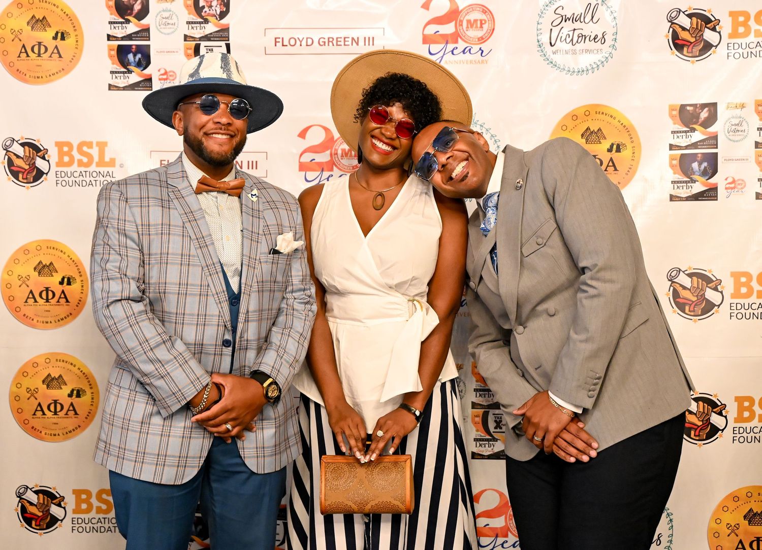 Three people in stylish attire pose together at an event with branded step-and-repeat backdrop.