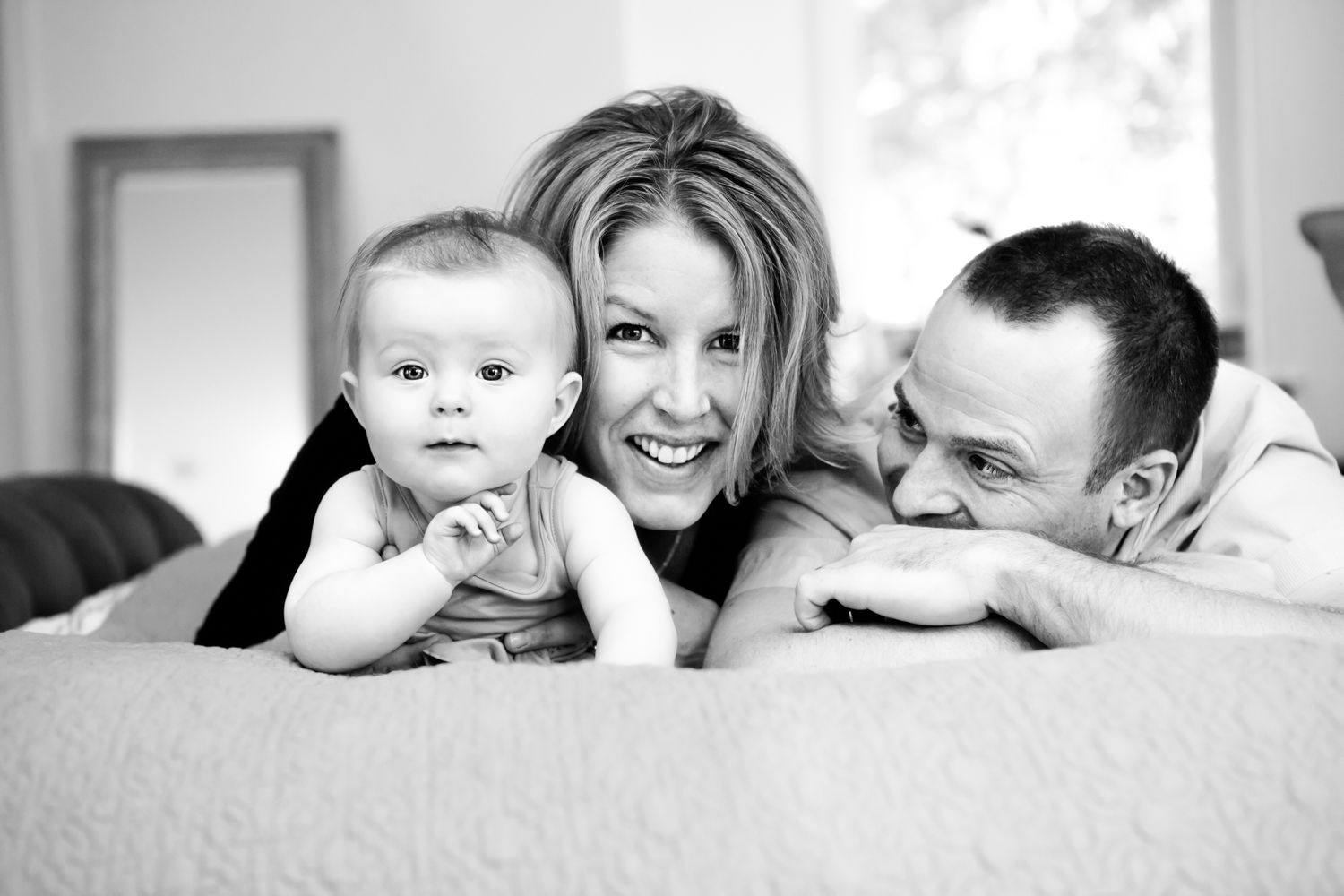 Black and white family portrait of smiling parents lying down with their baby on a soft white surface.