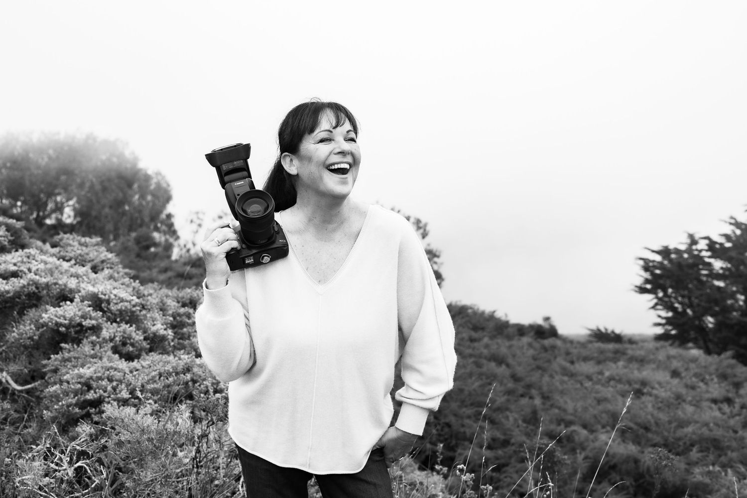 A laughing woman holds a professional camera outdoors among lush greenery in a black and white photo.