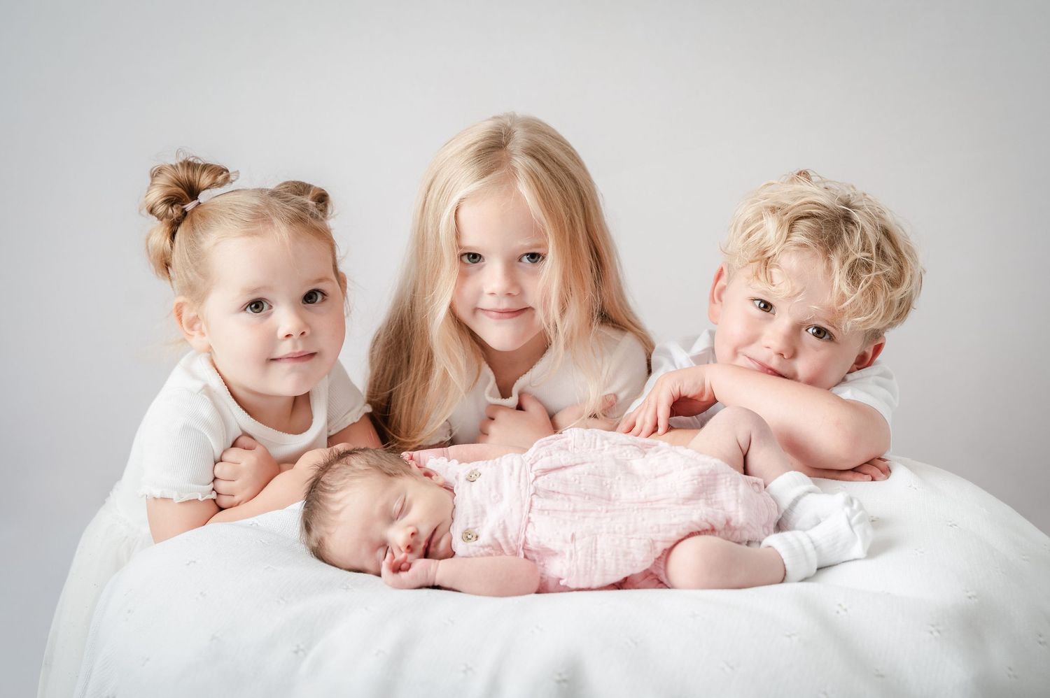 A series of tender newborn photography portraits showing siblings bonding with a baby on white bedding.