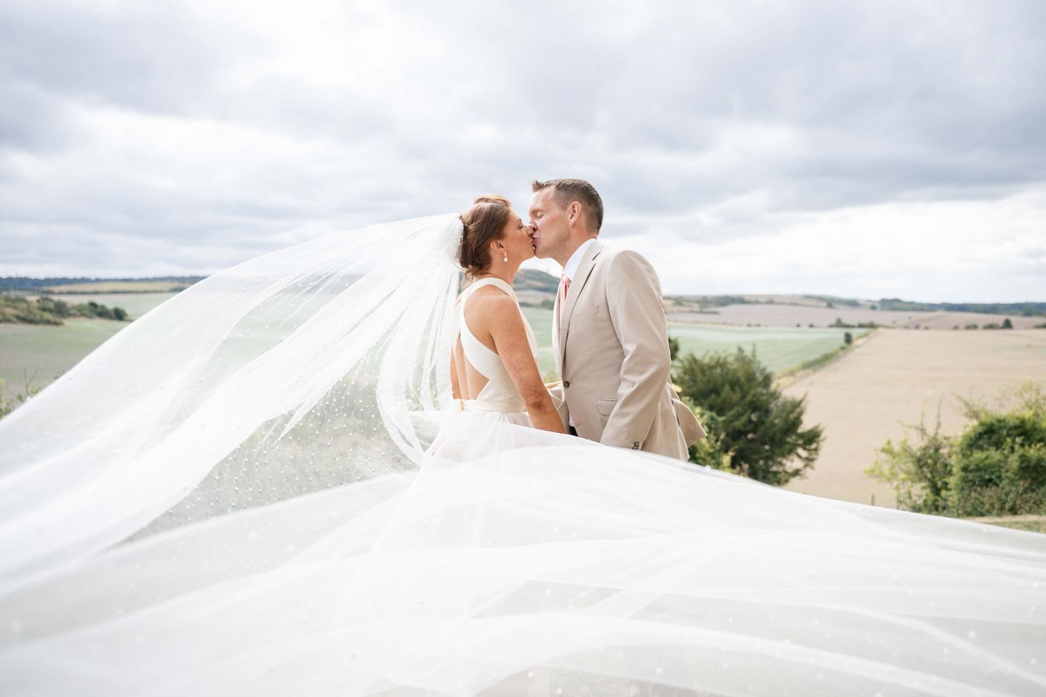 A romantic couple shares a kiss outdoors with a flowing white veil blowing dramatically in the wind.