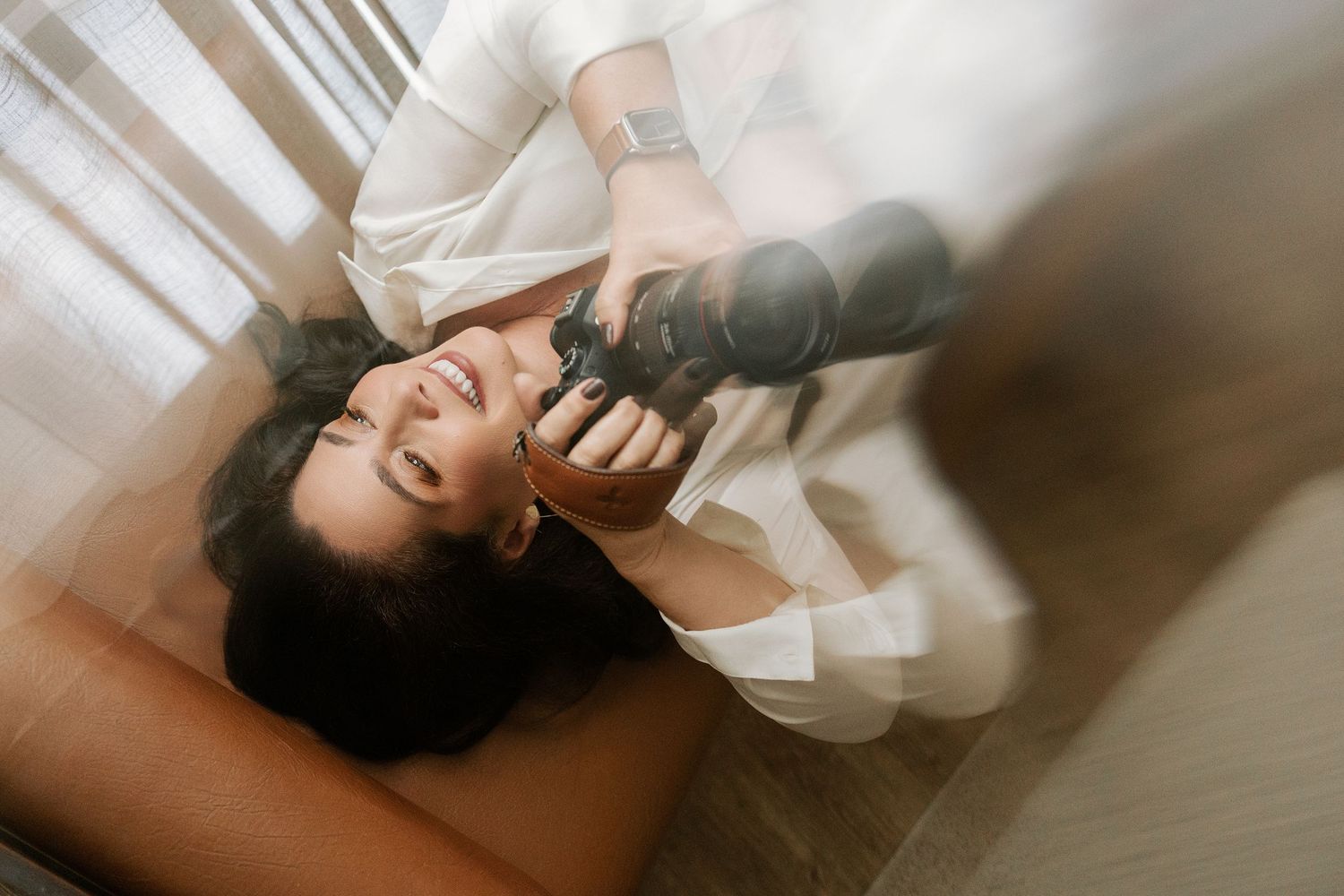 A smiling woman in white holds a camera while lying down, captured from an overhead angle in soft natural light.