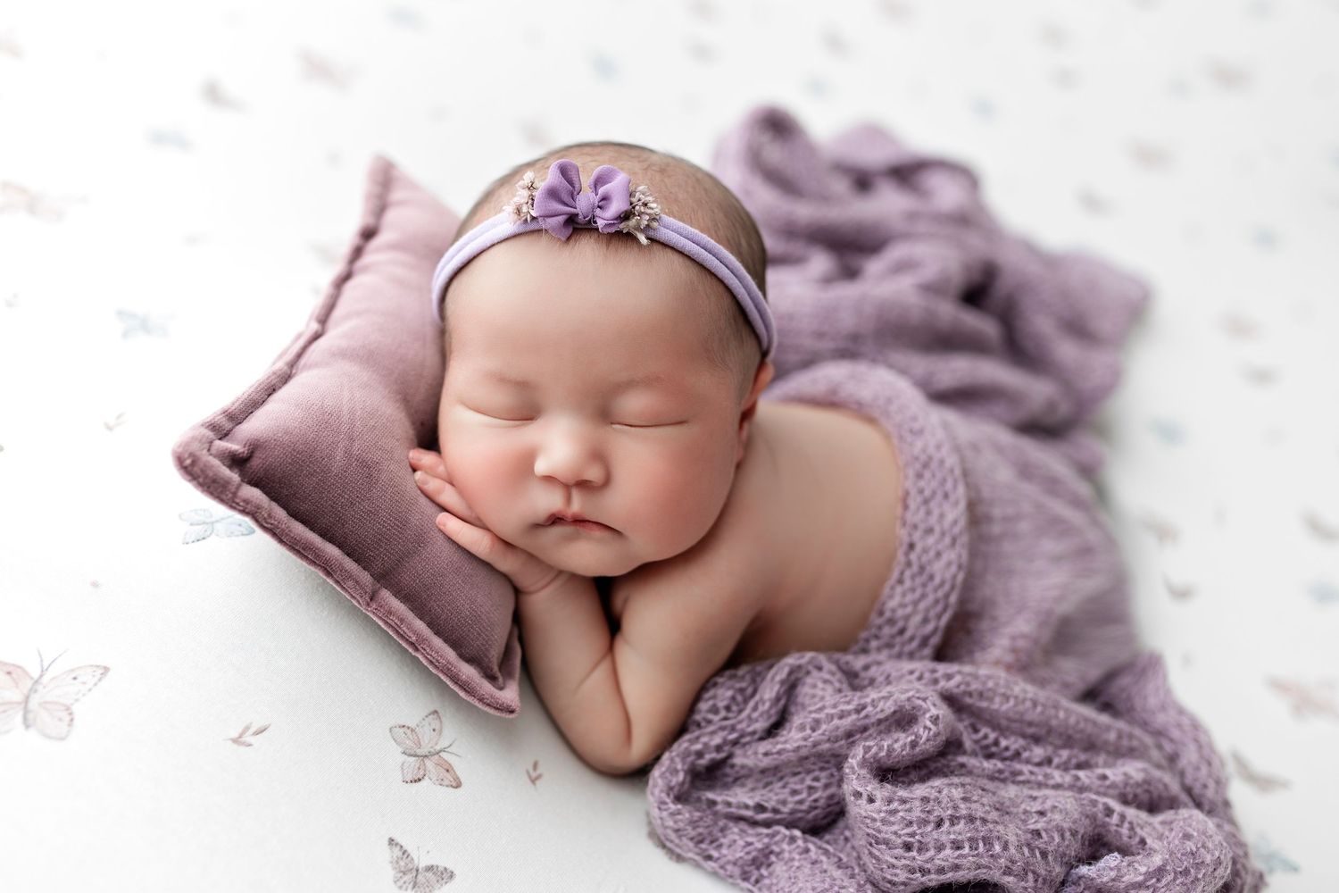 Newborn baby sleeping peacefully on soft purple blanket with delicate flower headband and dreamy backdrop.