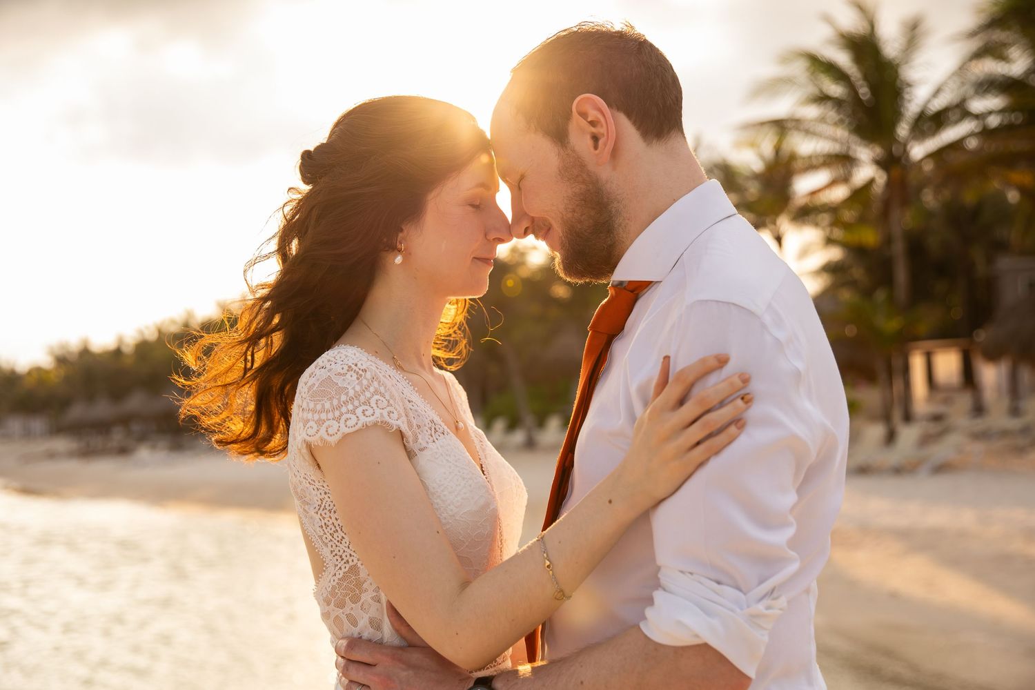 A romantic sunset beach portrait of a couple in wedding attire embracing with palm trees in background.
