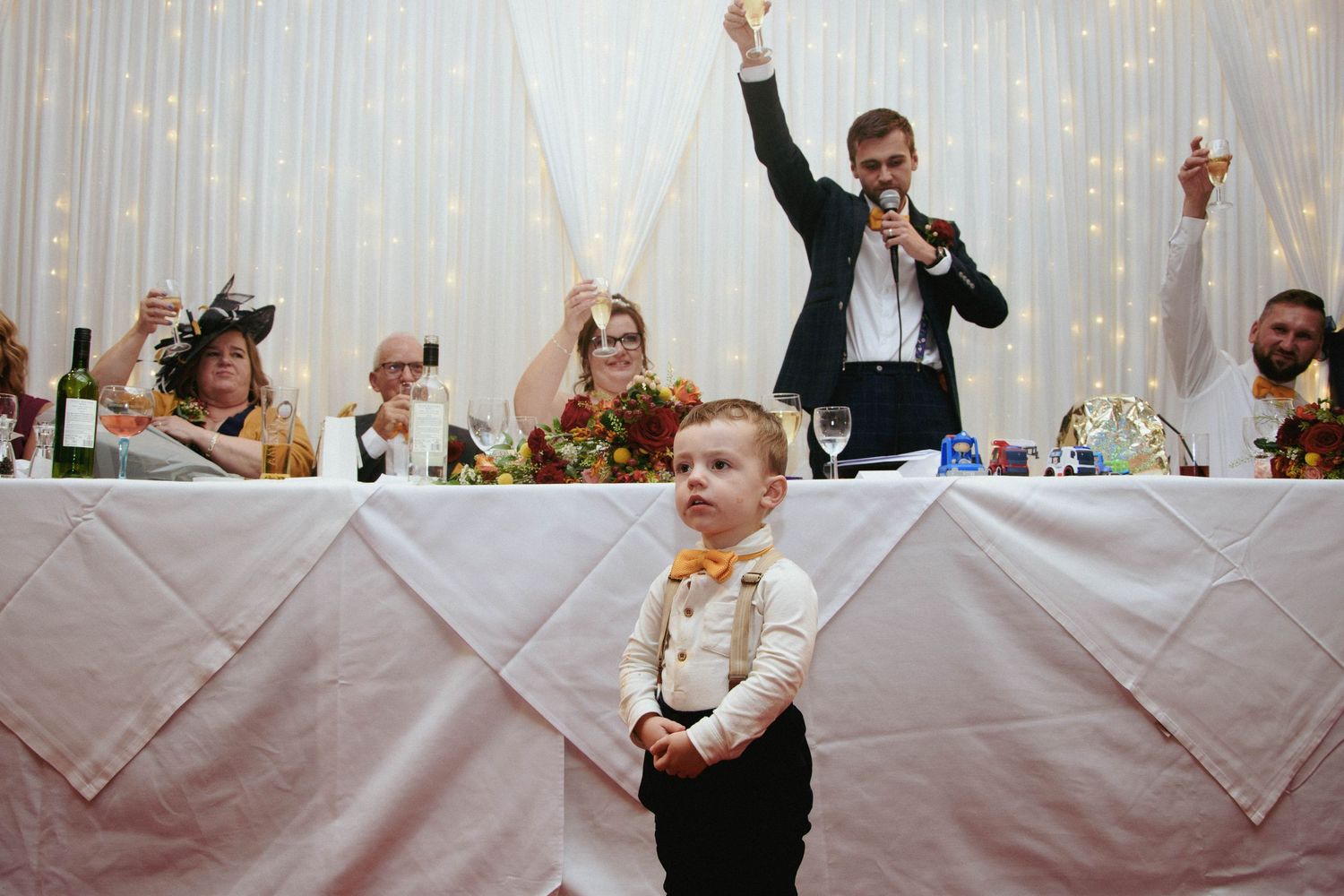 A young boy in suspenders and an orange bow tie stands before a wedding head table as a man gives a speech with a microphone.