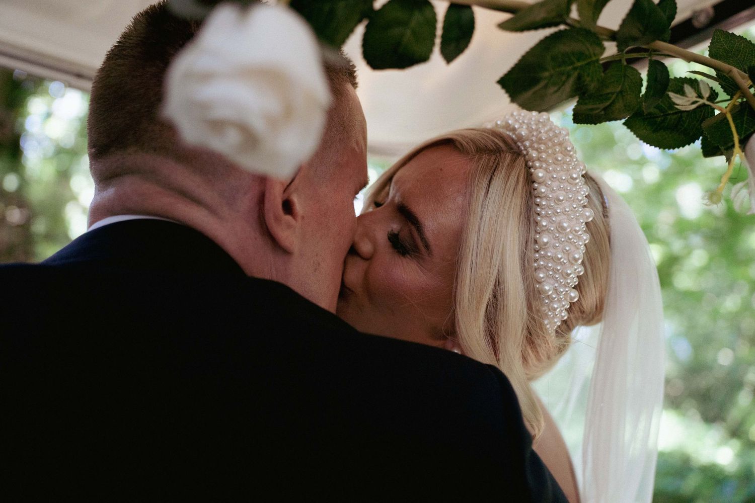 A bride in a pearl headpiece and veil kisses her groom under a leafy canopy decorated with white flowers at a wedding ceremony.