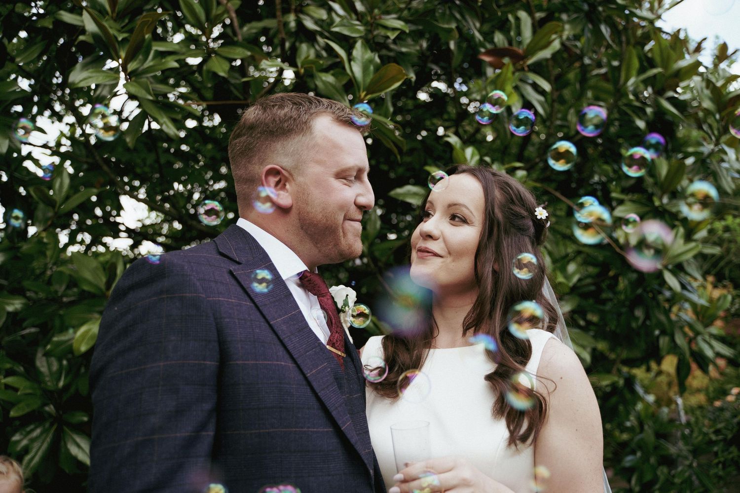Bride and groom in a tweed suit smile at each other among floating soap bubbles in front of leafy green foliage.