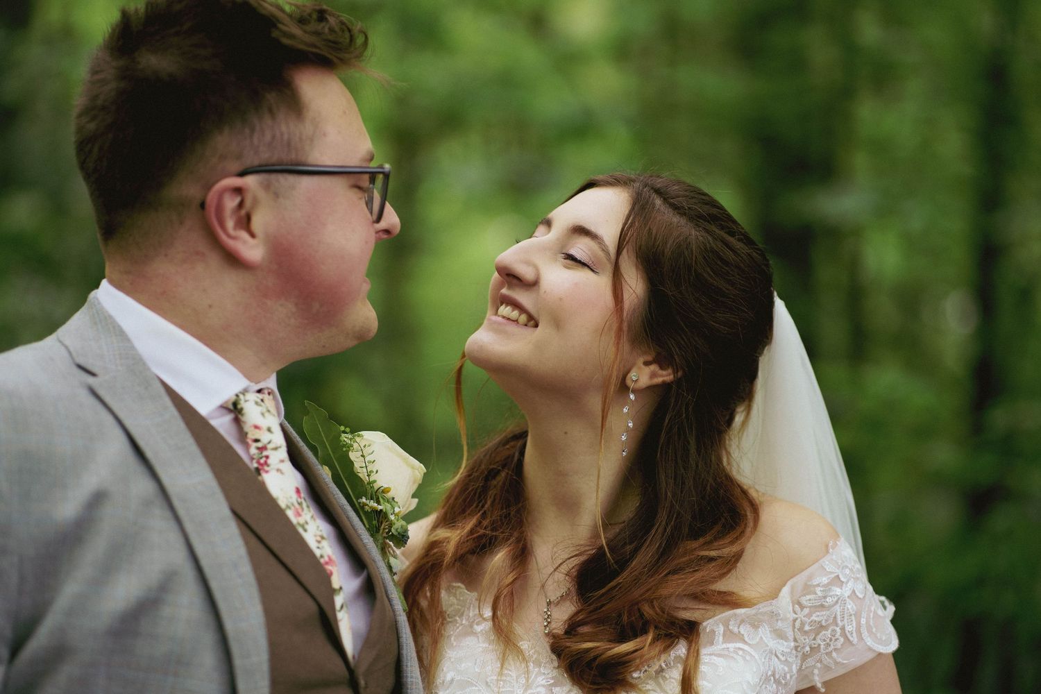 A bride in a lace dress and veil gazes up at her smiling groom in a grey suit and floral tie against a green forest backdrop.