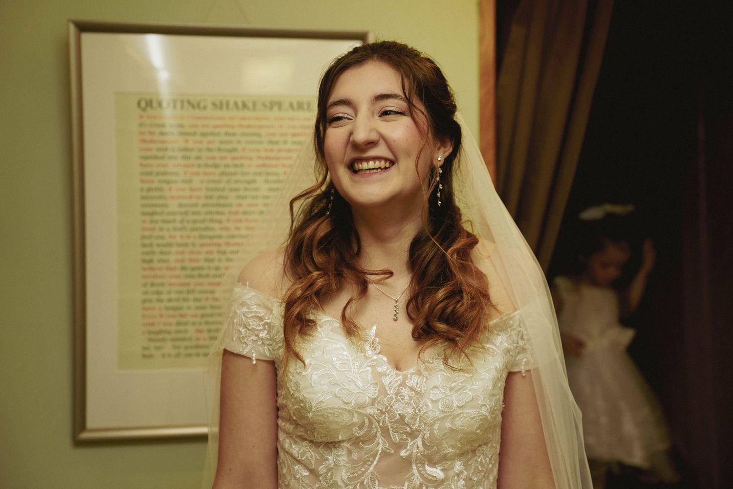 A smiling bride in a lace wedding dress and veil laughs happily indoors while a flower girl stands in the background.