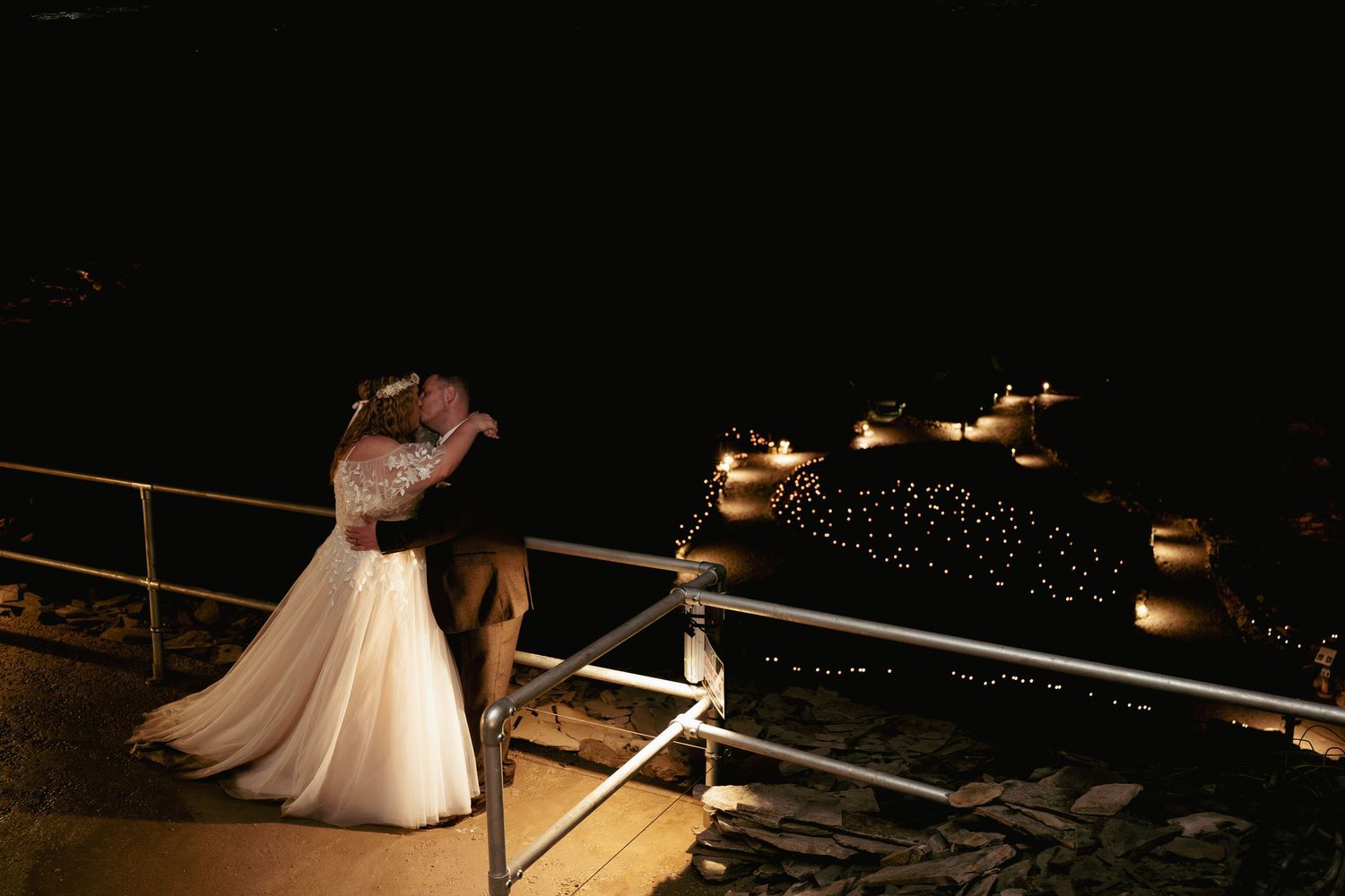 A bride in a white gown and a groom embrace on a railed terrace at night beside softly glowing candles.