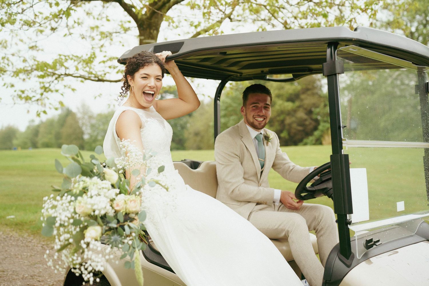 A joyful bride in a white gown laughs on the back of a golf cart while the groom in a tan suit sits at the wheel.