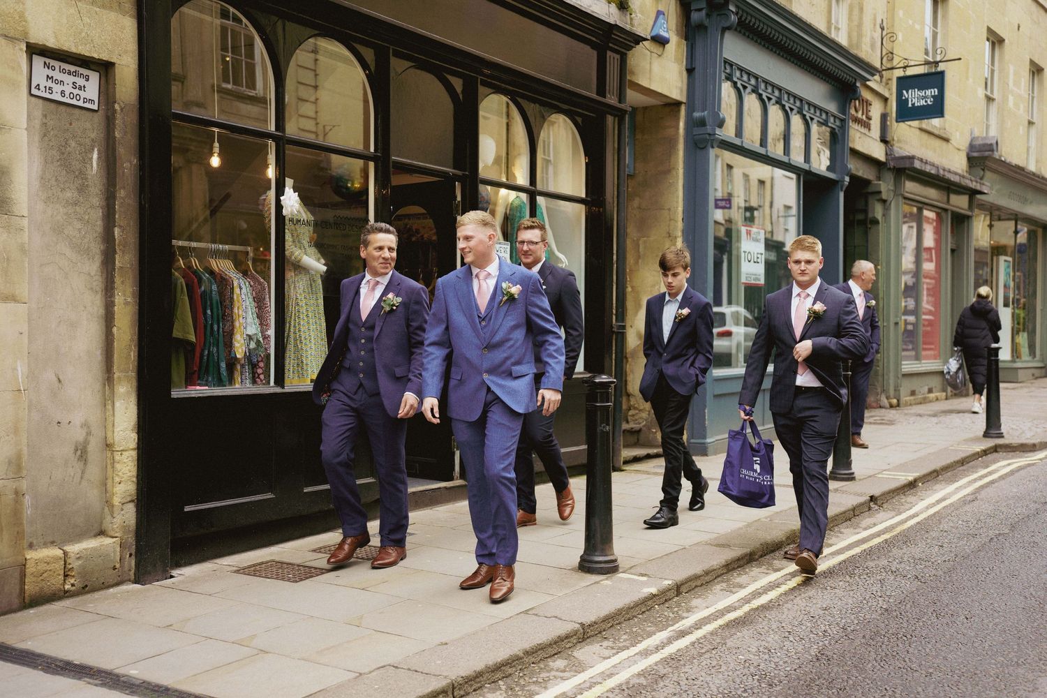 Groomsmen in matching blue suits walk together down a cobblestone street past shop windows on their wedding day.