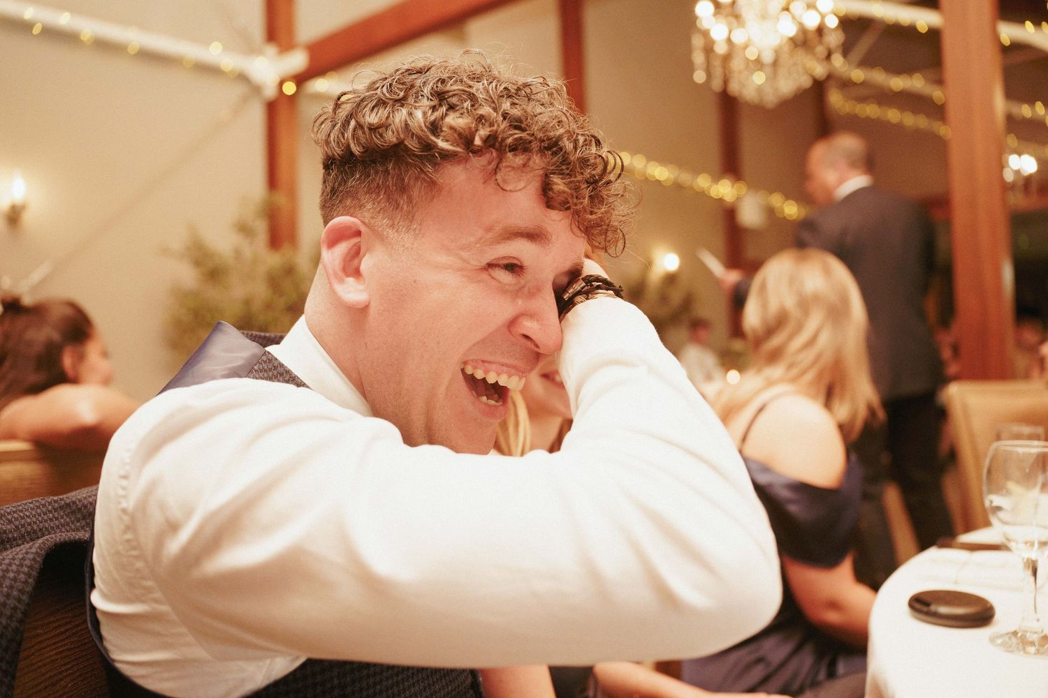 A laughing man in a white shirt and vest rests his head on his hand during a lively wedding reception dinner.