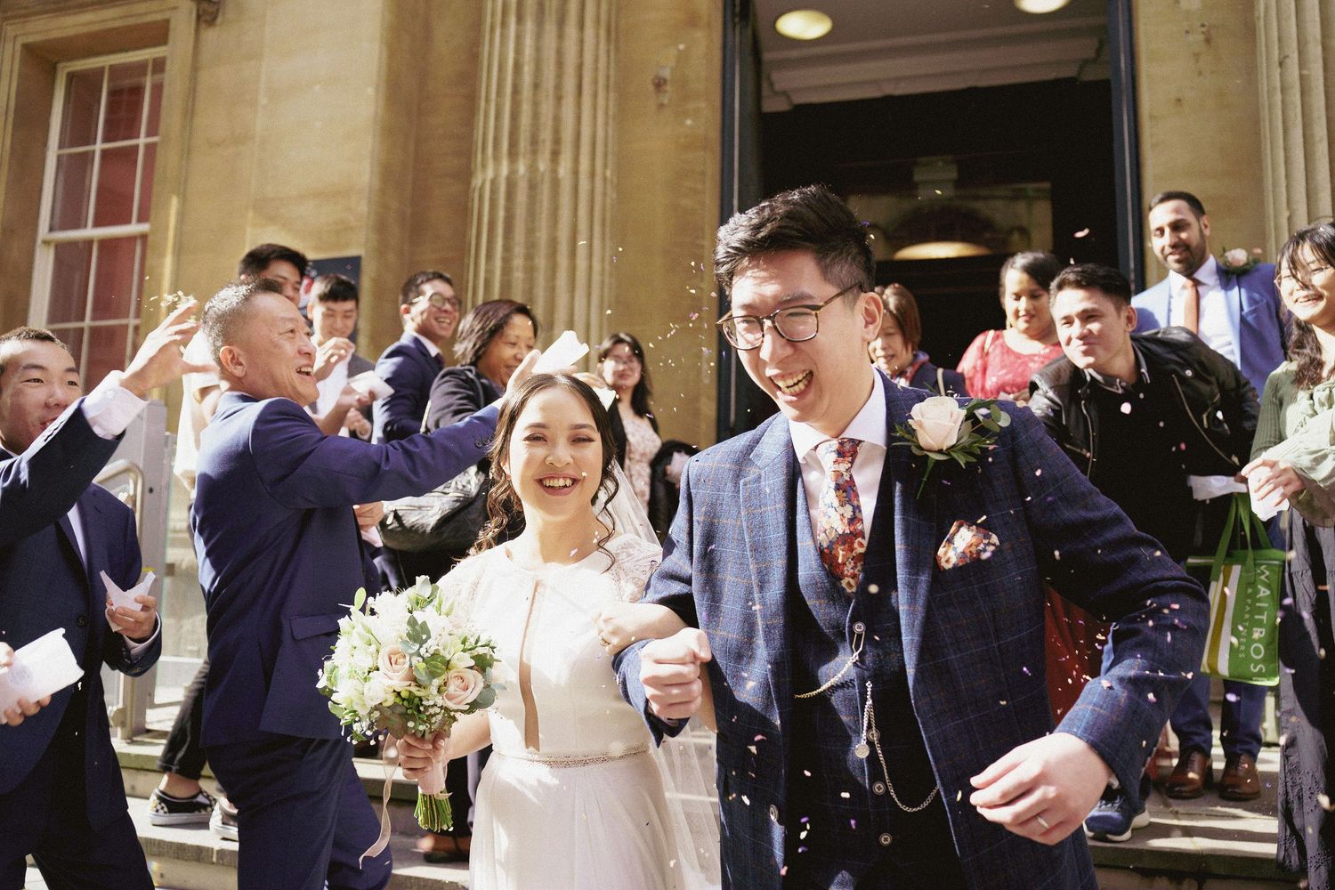 A smiling bride holding a bouquet and a groom in a navy suit leave a ceremony as guests cheer around them.