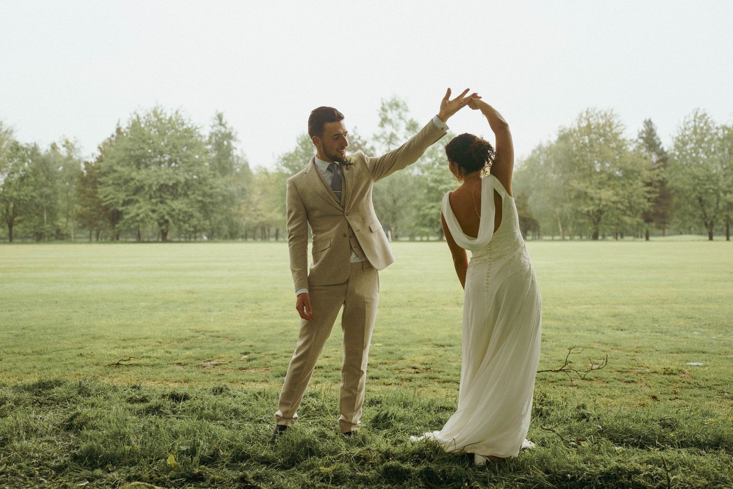 A bride and groom in a tan suit dance and twirl together on a misty green lawn surrounded by trees.