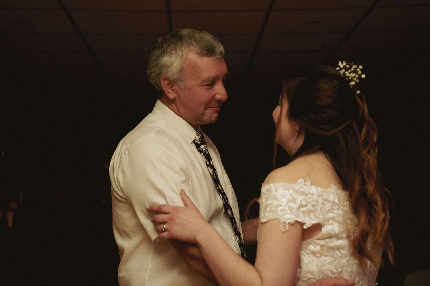 A bride in a lace wedding dress shares a dance with a smiling older man in a white shirt and tie in a dimly lit reception room.