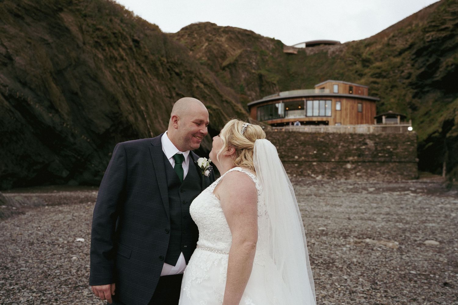 Smiling bride in a veil and groom in a tweed waistcoat stand face to face on a pebble beach with cliffs and a coastal building behind.